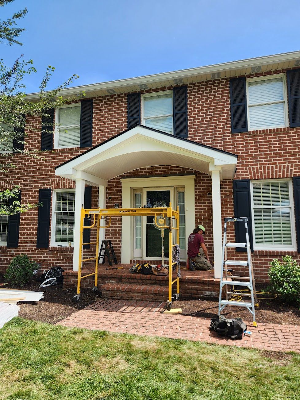 A person in a red shirt working on the front porch of a brick house with black shutters and a yellow scaffolding unit.
