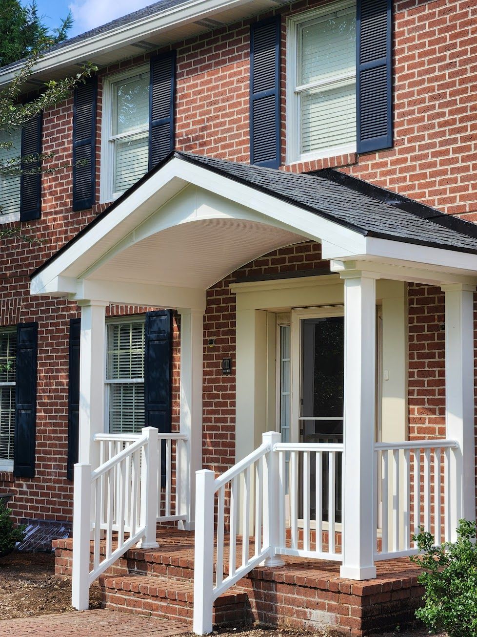 Red brick home entrance with a white gabled porch, arched ceiling, and white railings over brick steps.