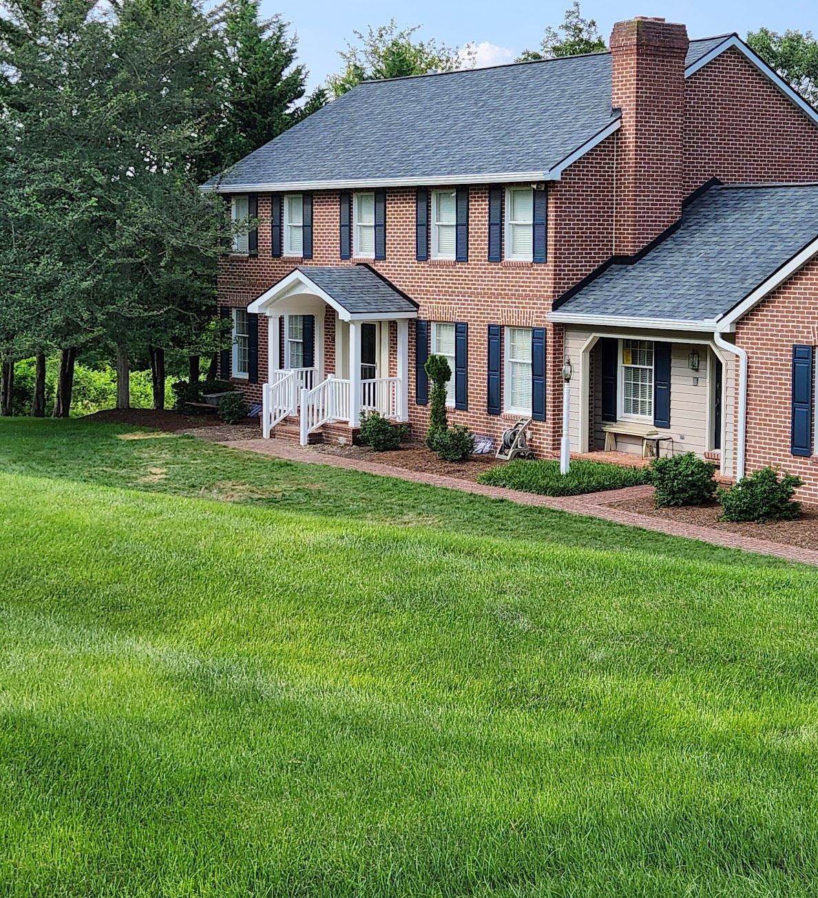 A two-story red brick house with dark shutters and a grey roof, situated on a grassy hill with surrounding trees.