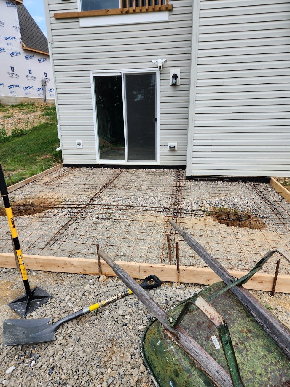 A construction site for a concrete patio with wire mesh reinforcement and wooden forms set against a residential house.