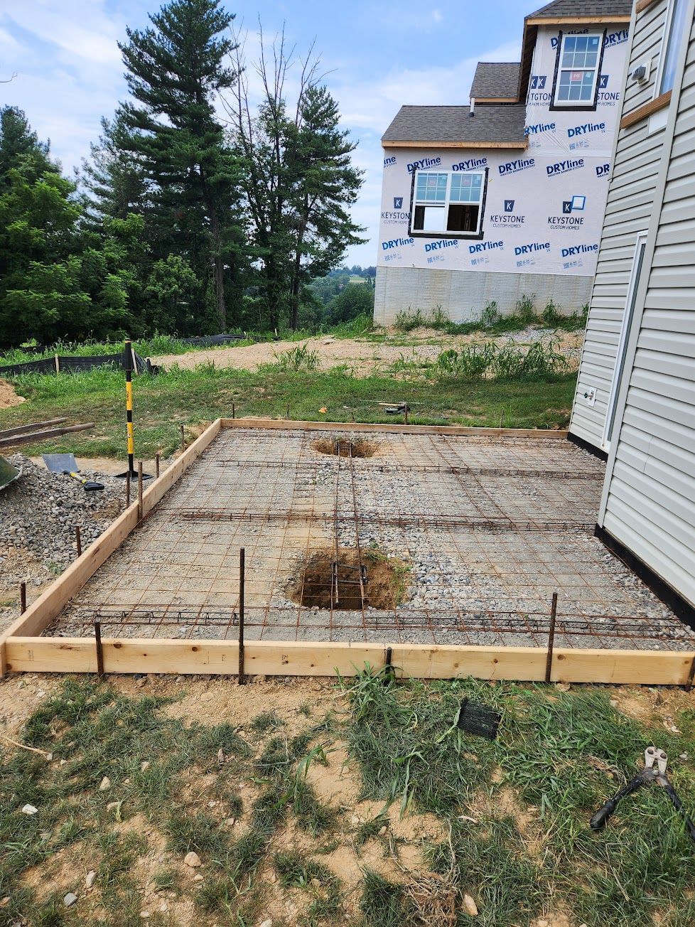 A wooden frame for a concrete slab foundation sits on gravel, with steel rebar visible, next to a house under construction.