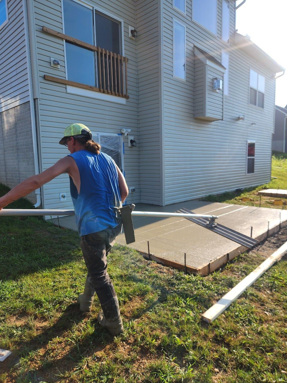 A person in a blue tank top uses a long-handled rake to smooth freshly poured concrete for a new patio beside a house.