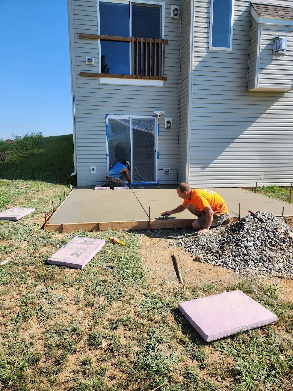 Two workers finishing a freshly poured concrete patio slab outside the back of a house.