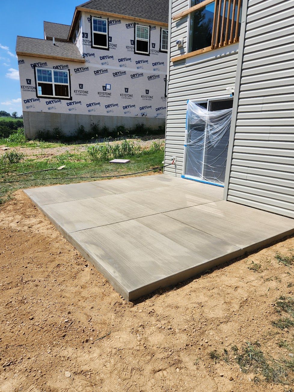 A newly poured rectangular concrete patio sits beside a house with beige siding and a plastic-covered sliding door.