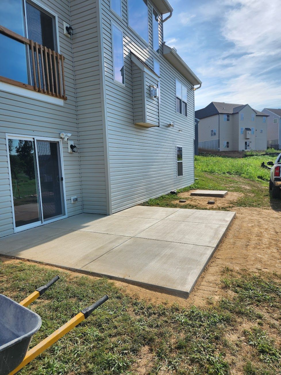 A newly poured square concrete patio sits against the side of a light-gray vinyl-sided house on a sunny day.
