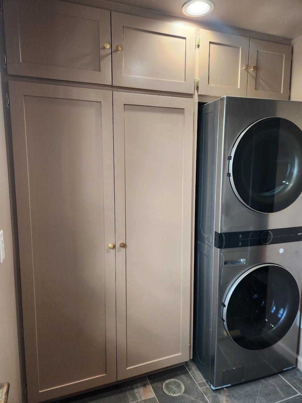A stacked stainless steel washer and dryer next to tall, light-brown pantry cabinets in a home laundry room.