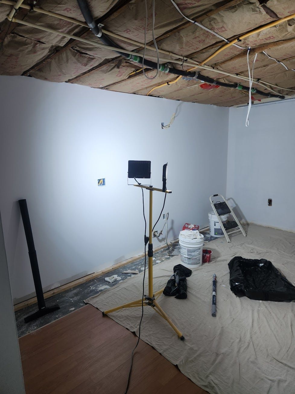 An unfinished room under renovation with exposed ceiling insulation, a work light on a tripod, and tools on the floor.