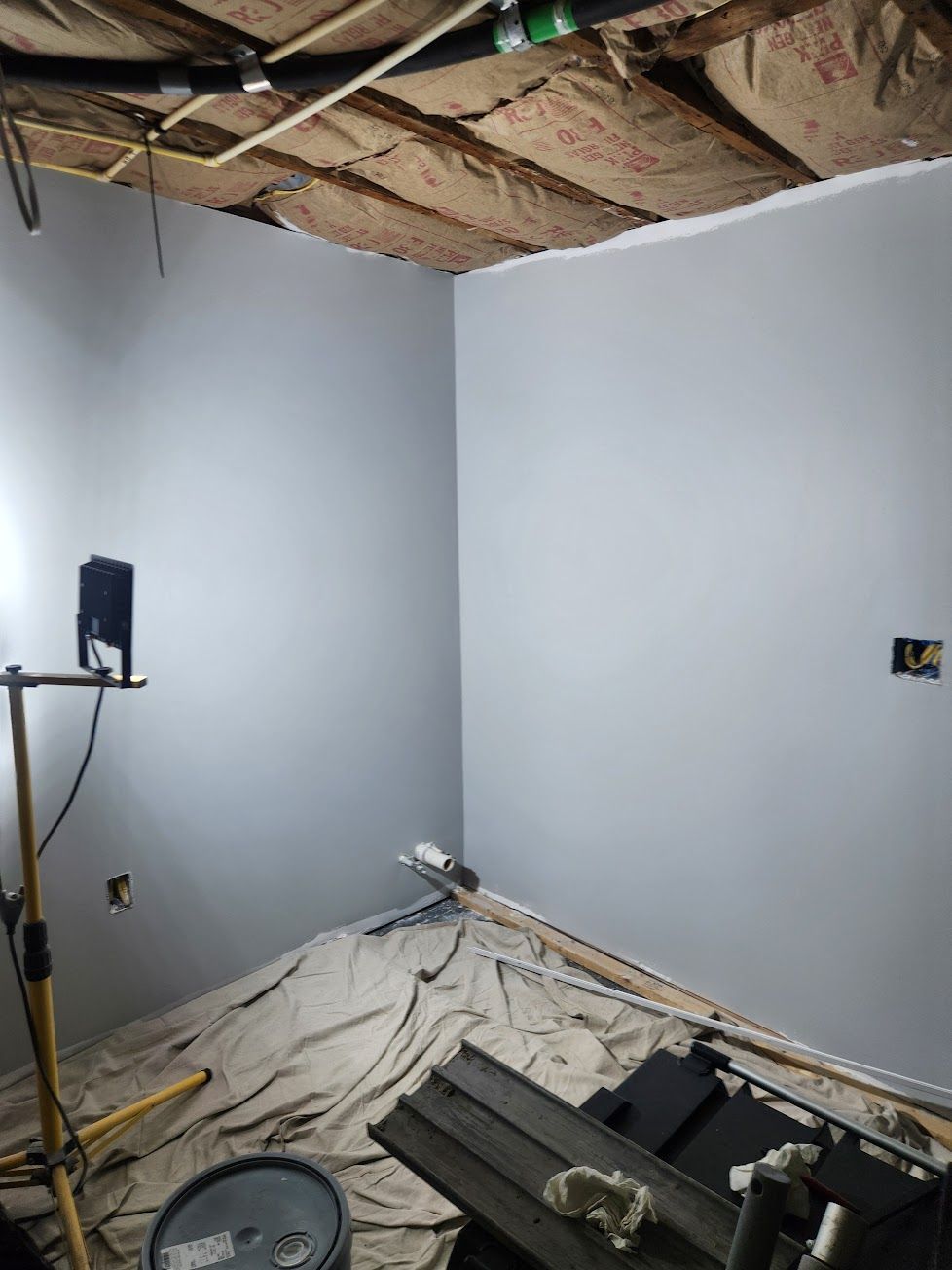 A room under construction with grey painted walls, exposed ceiling insulation, a work light, and a drop cloth on the floor.