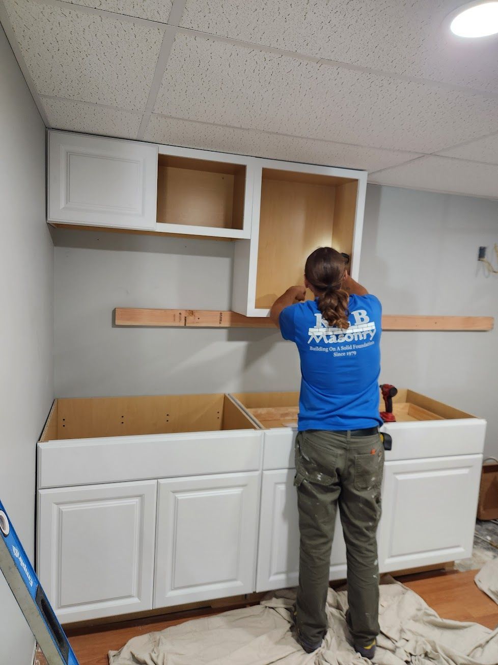 A person in a blue shirt installs white cabinets onto a wall above base cabinets in a room with light grey walls.