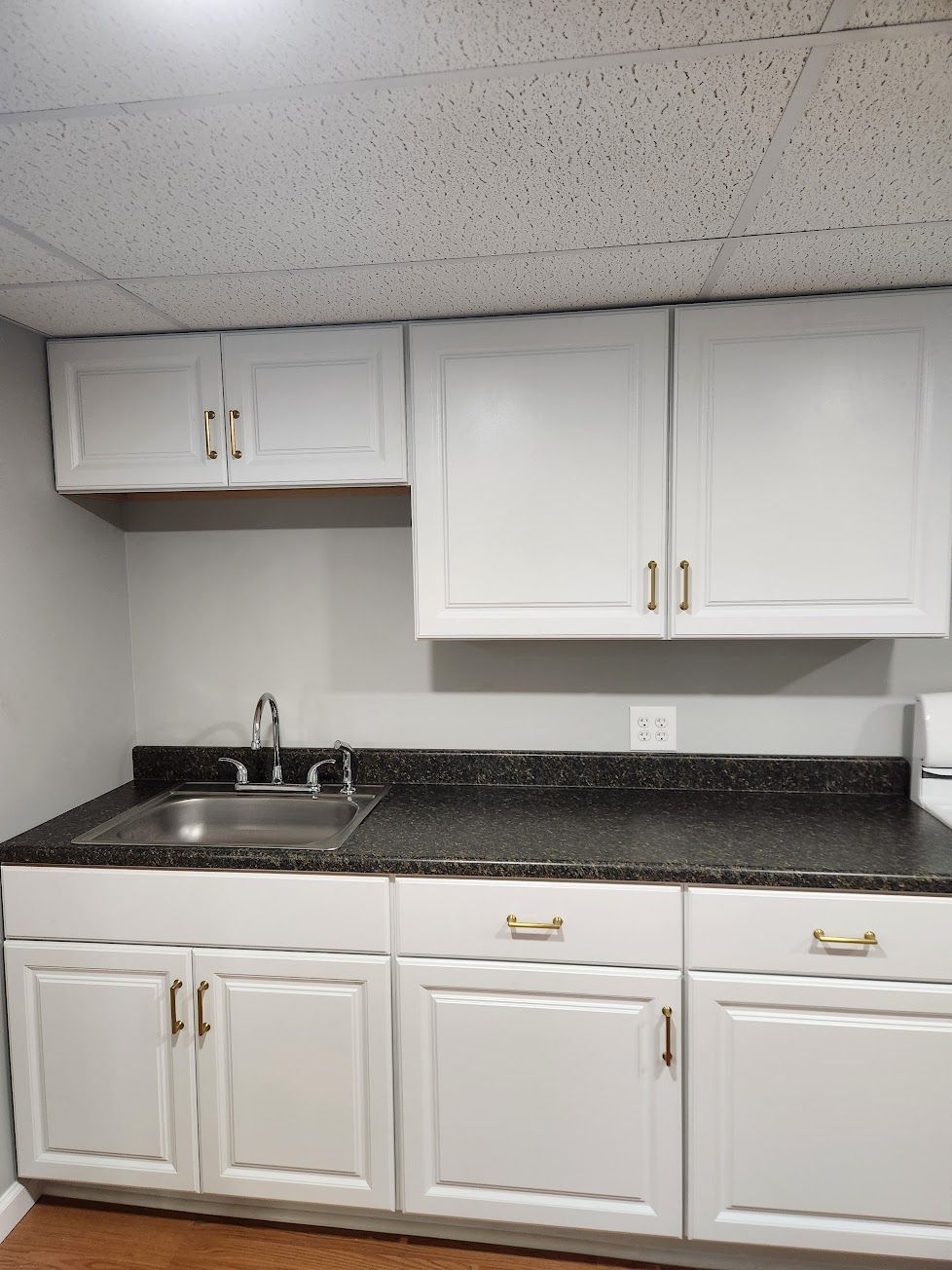A kitchen kitchenette with white cabinets, a black countertop, and a stainless steel sink against a plain light-gray wall.