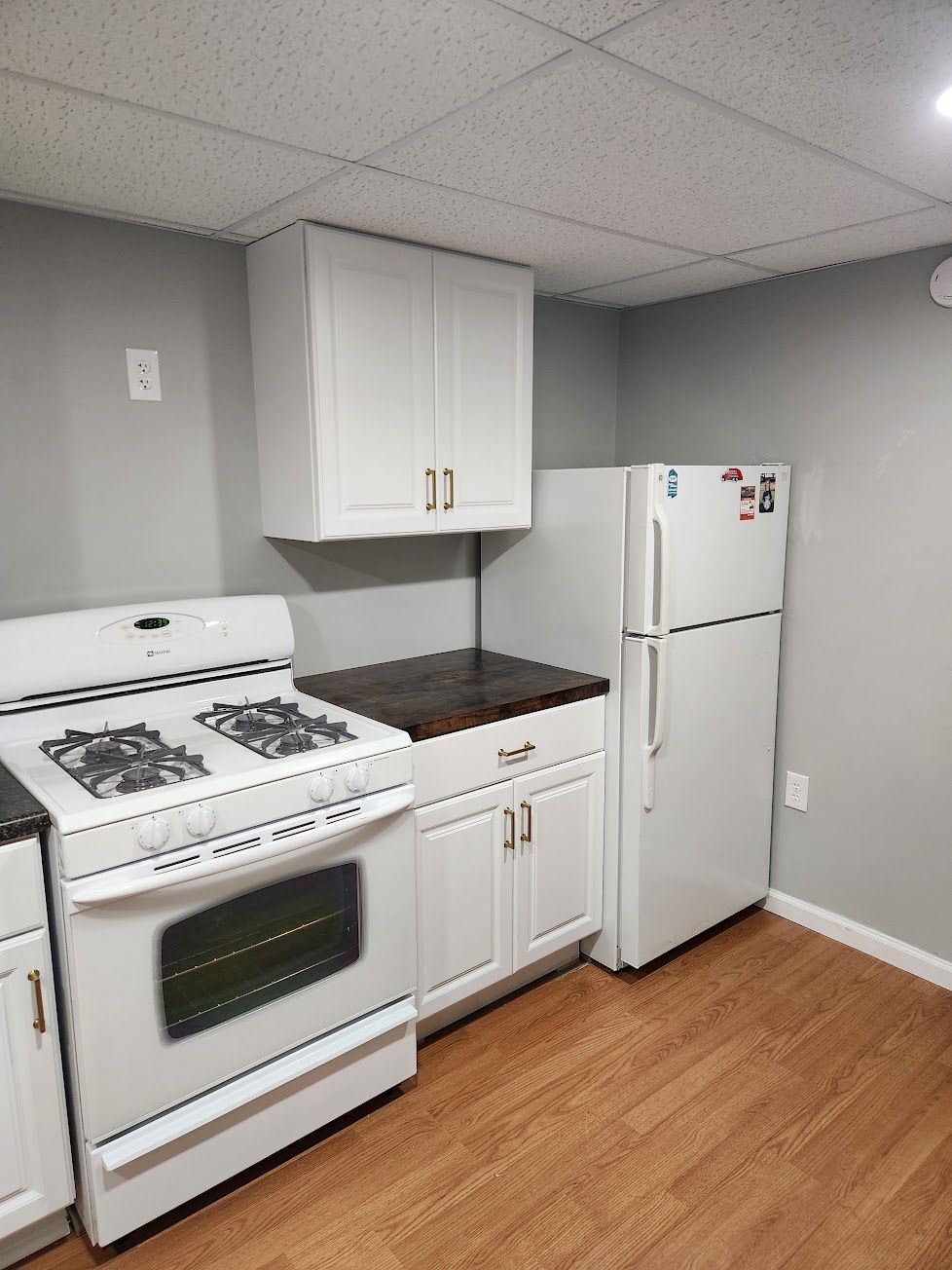 A white gas stove, a refrigerator, and a kitchen cabinet with a dark wood countertop in a room with grey walls.