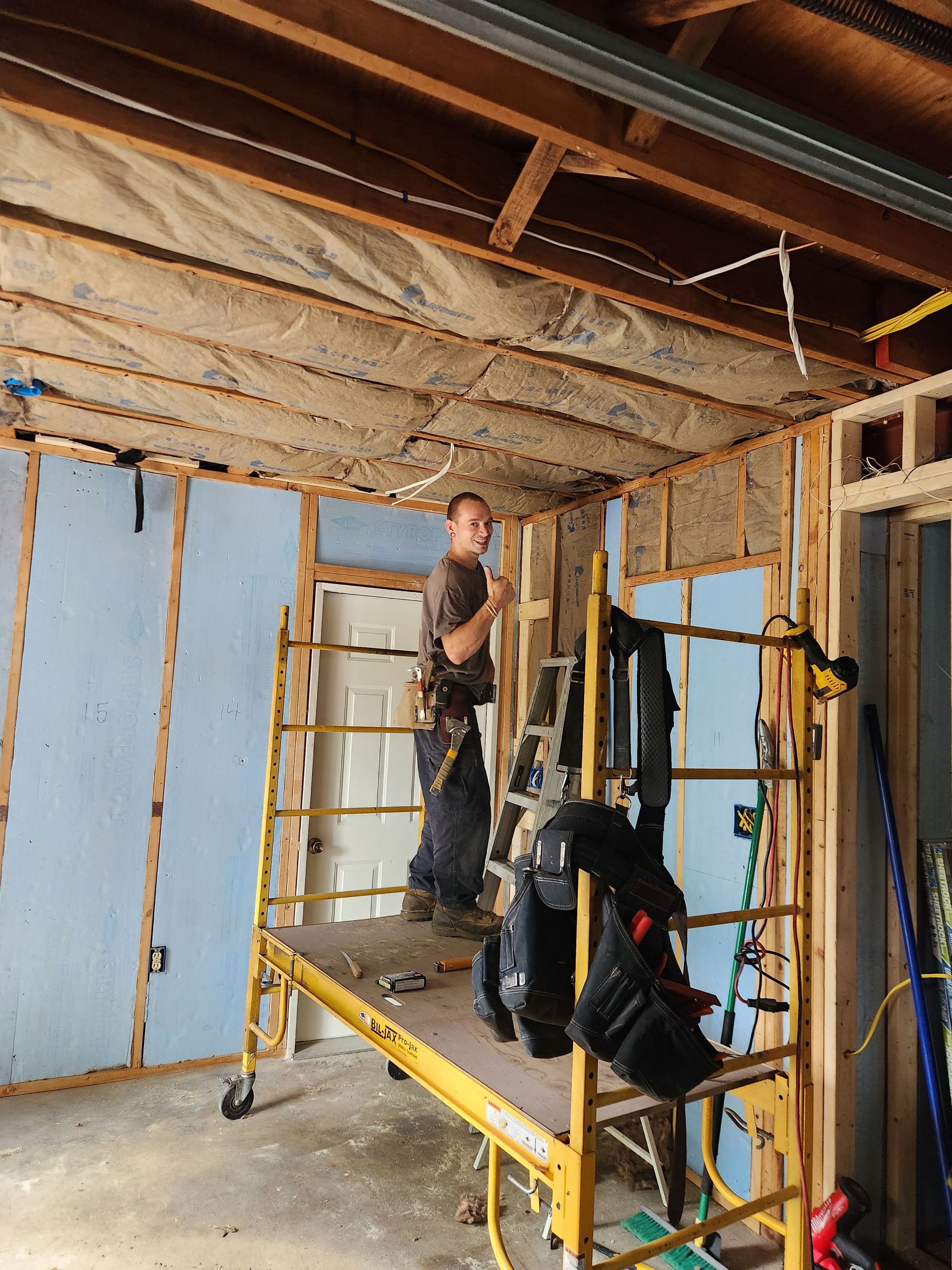 A person stands on yellow scaffolding, installing insulation in the unfinished wood-framed walls and ceiling of a room.