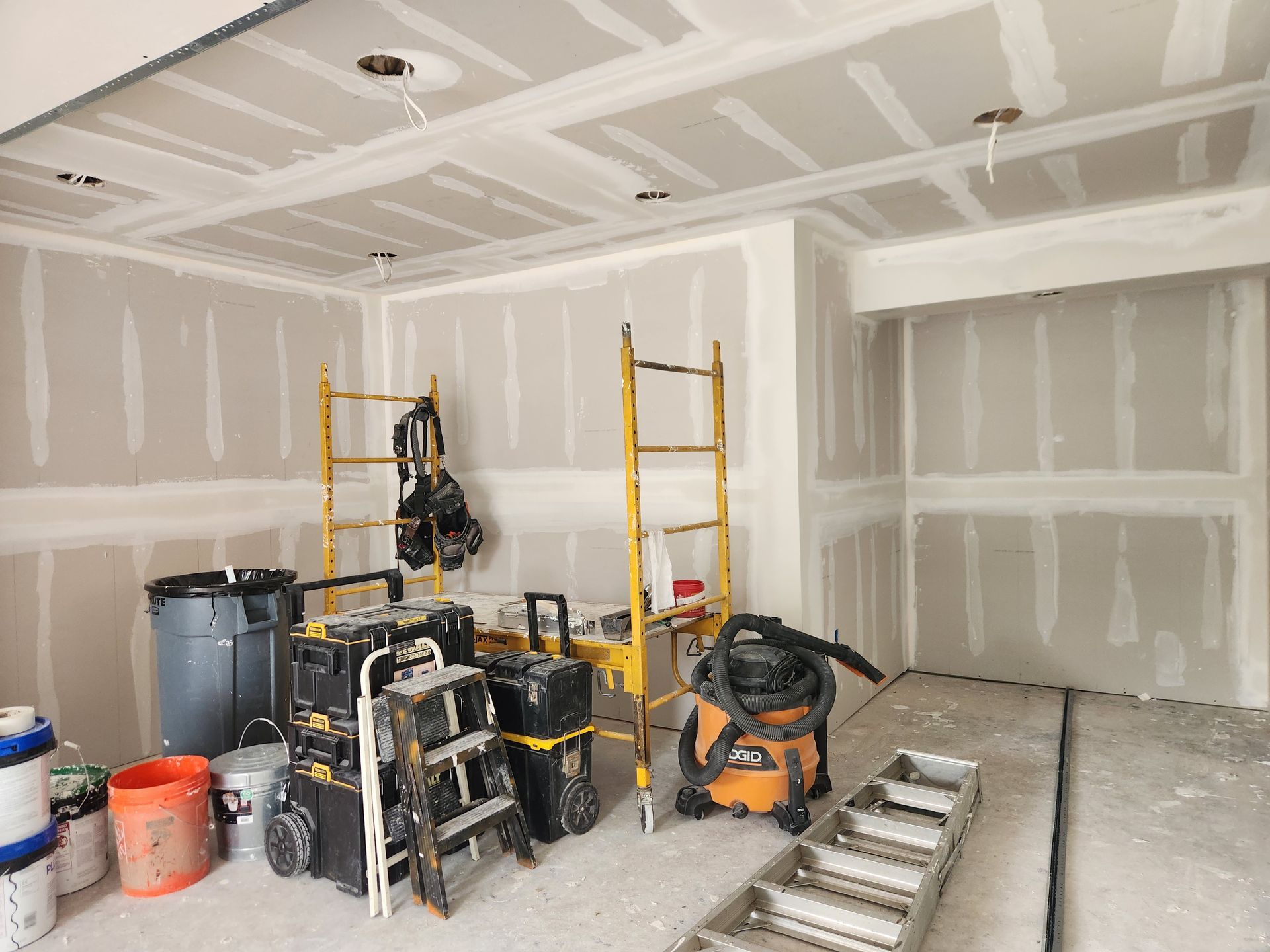 An unfinished room with drywall, a yellow scaffolding unit, a step ladder, construction tools, and containers on the floor.