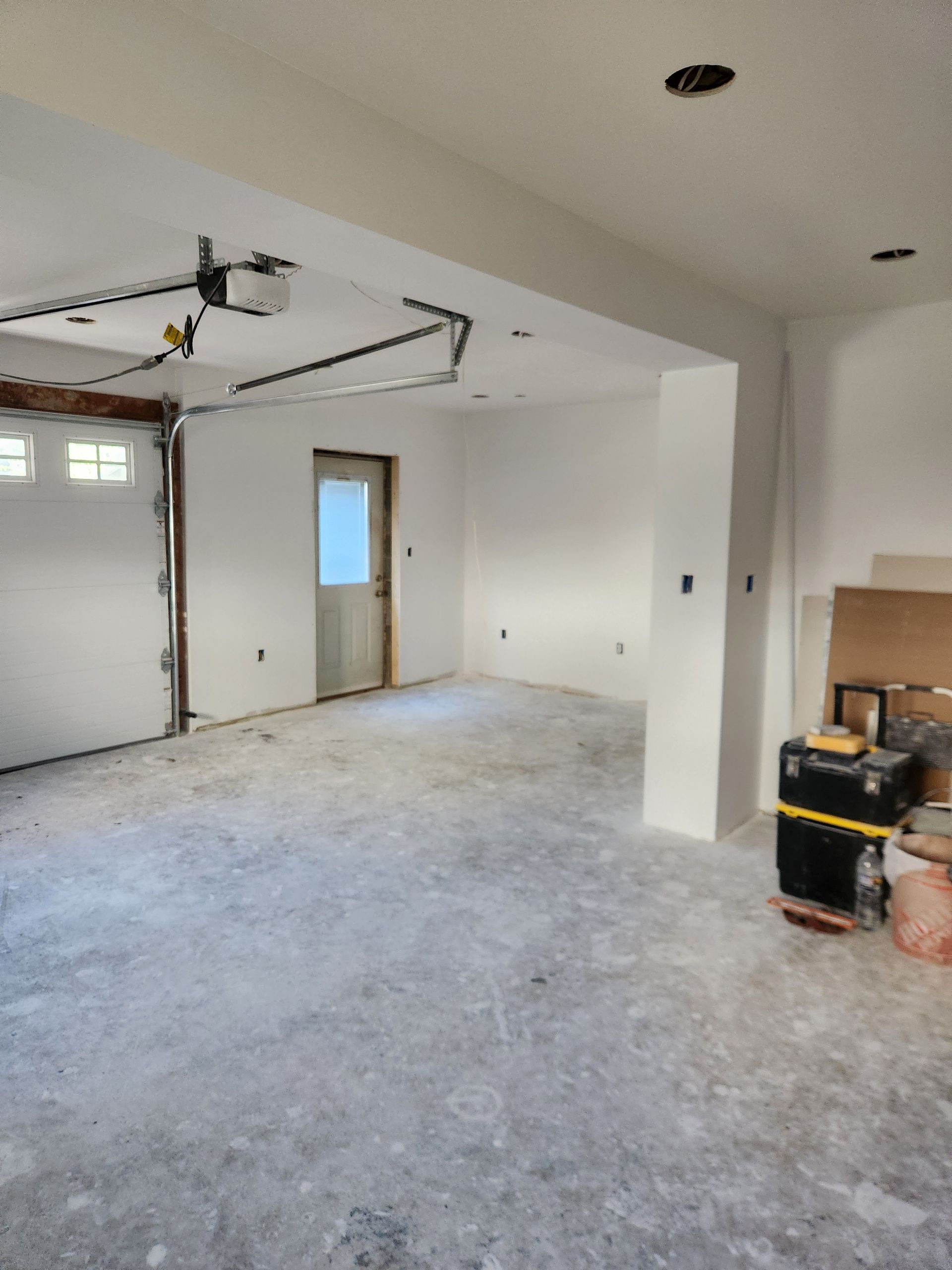 An unfinished garage with white walls, a gray concrete floor, a door leading outside, and a garage door opener.