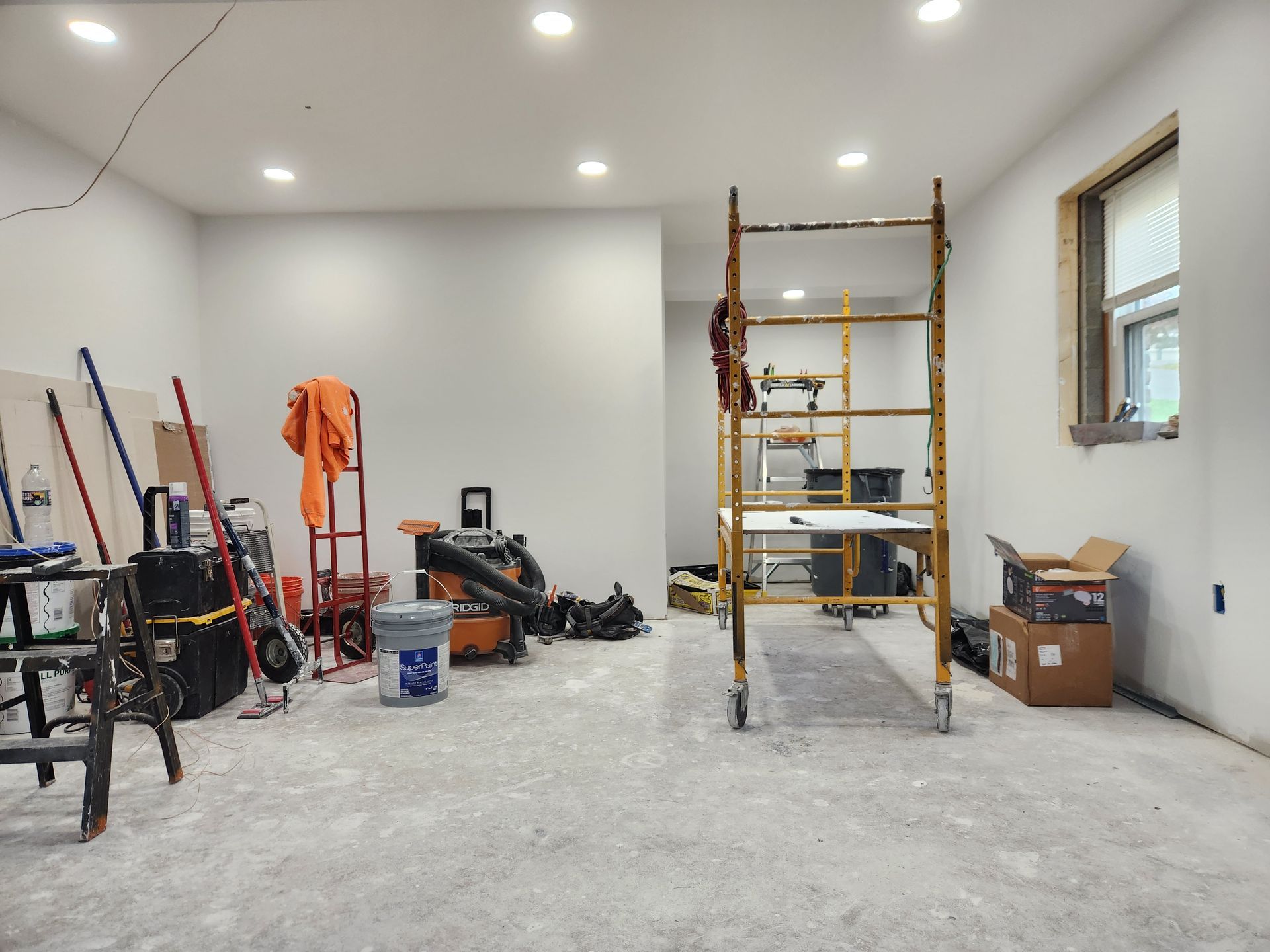 A room under construction with tools, a red ladder, scaffolding, and moving boxes on a concrete floor.