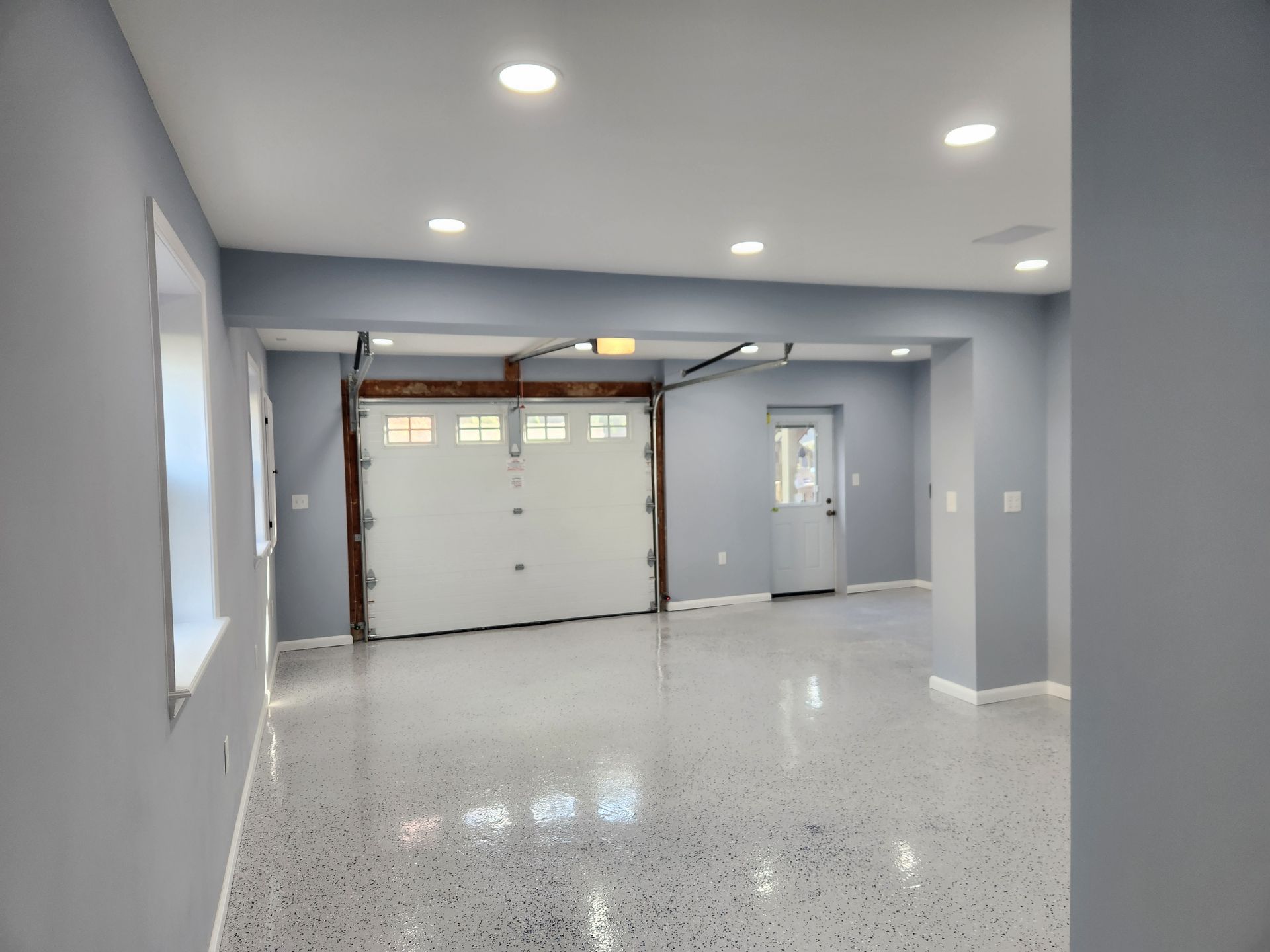 A modern garage with light blue walls, a white sectional door, and a speckled gray epoxy floor under recessed lighting.