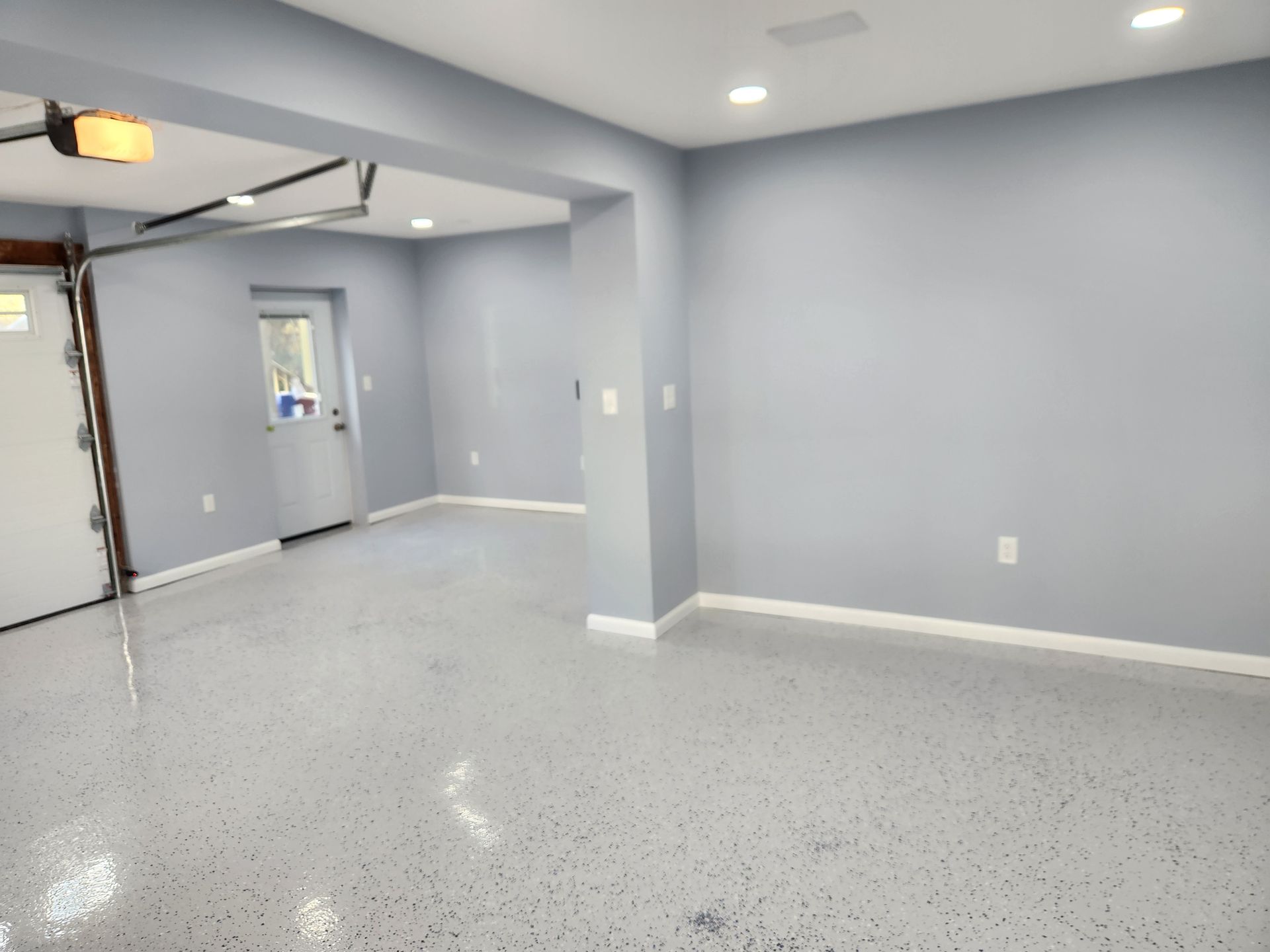 A modern garage interior with light gray walls, a speckled epoxy floor, a white door, and recessed ceiling lighting.