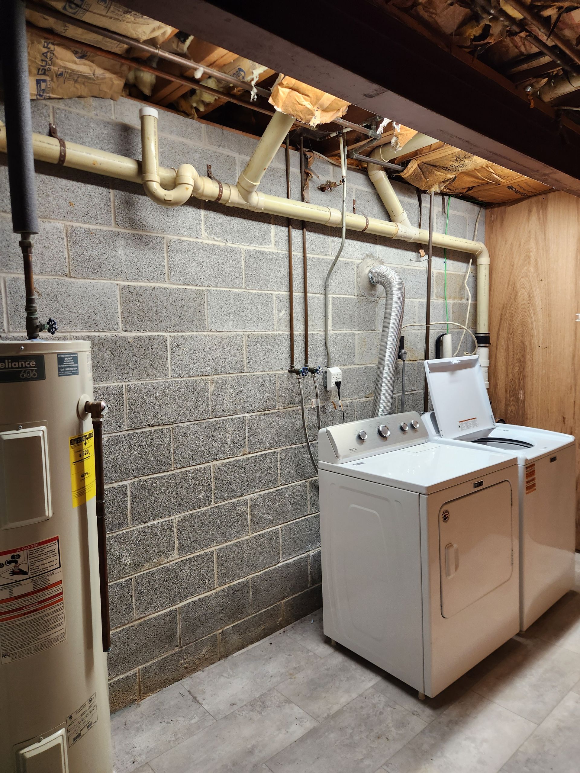 A basement laundry area with a water heater, a washer, a dryer, and exposed plumbing against a concrete block wall.