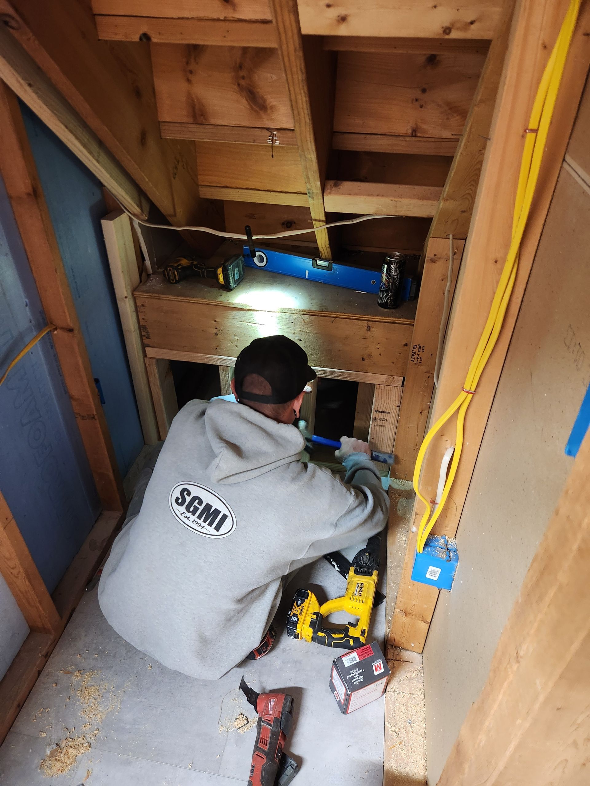 A person in a gray hoodie works in a wood-framed space, using a yellow power tool near stairs.