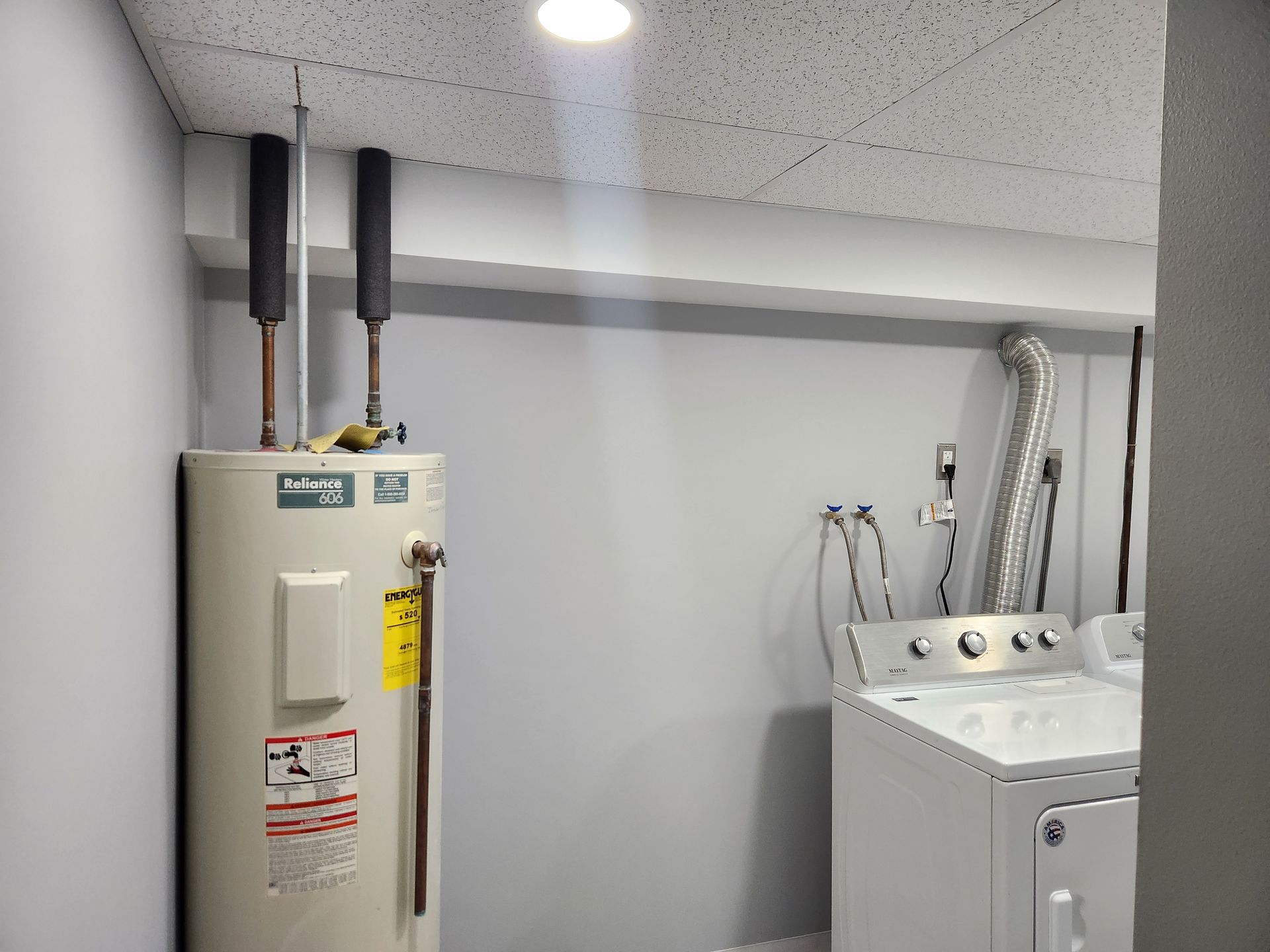 A water heater and a white clothes dryer side-by-side in a laundry room with gray walls and a drop ceiling.