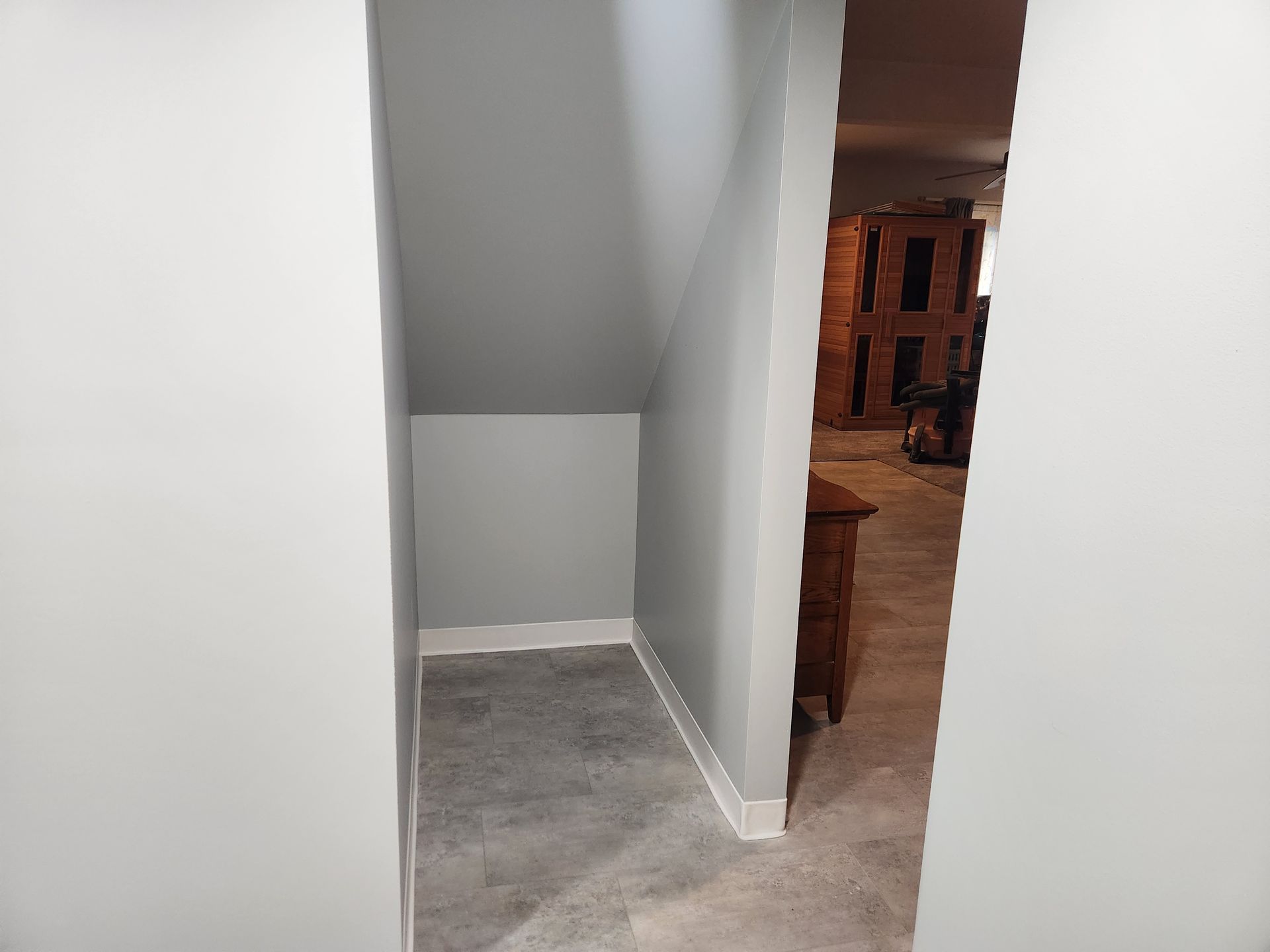 An alcove with gray walls and a gray floor under a staircase, next to an opening leading into a room with wood furniture.