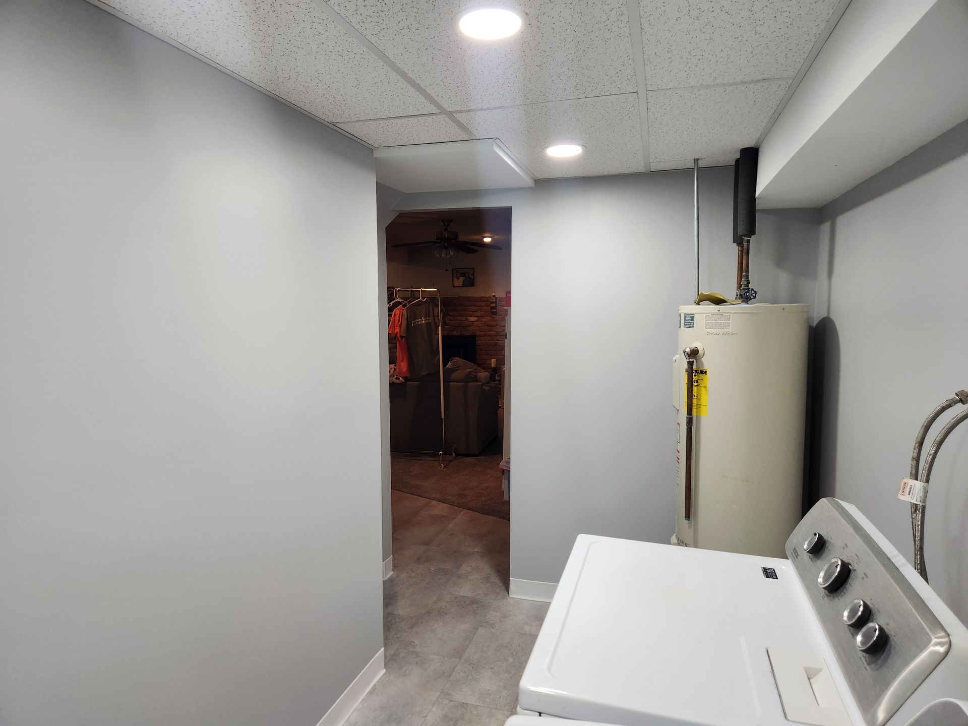 A utility room with a water heater and washing machine, featuring grey walls and a drop ceiling leading to another room.