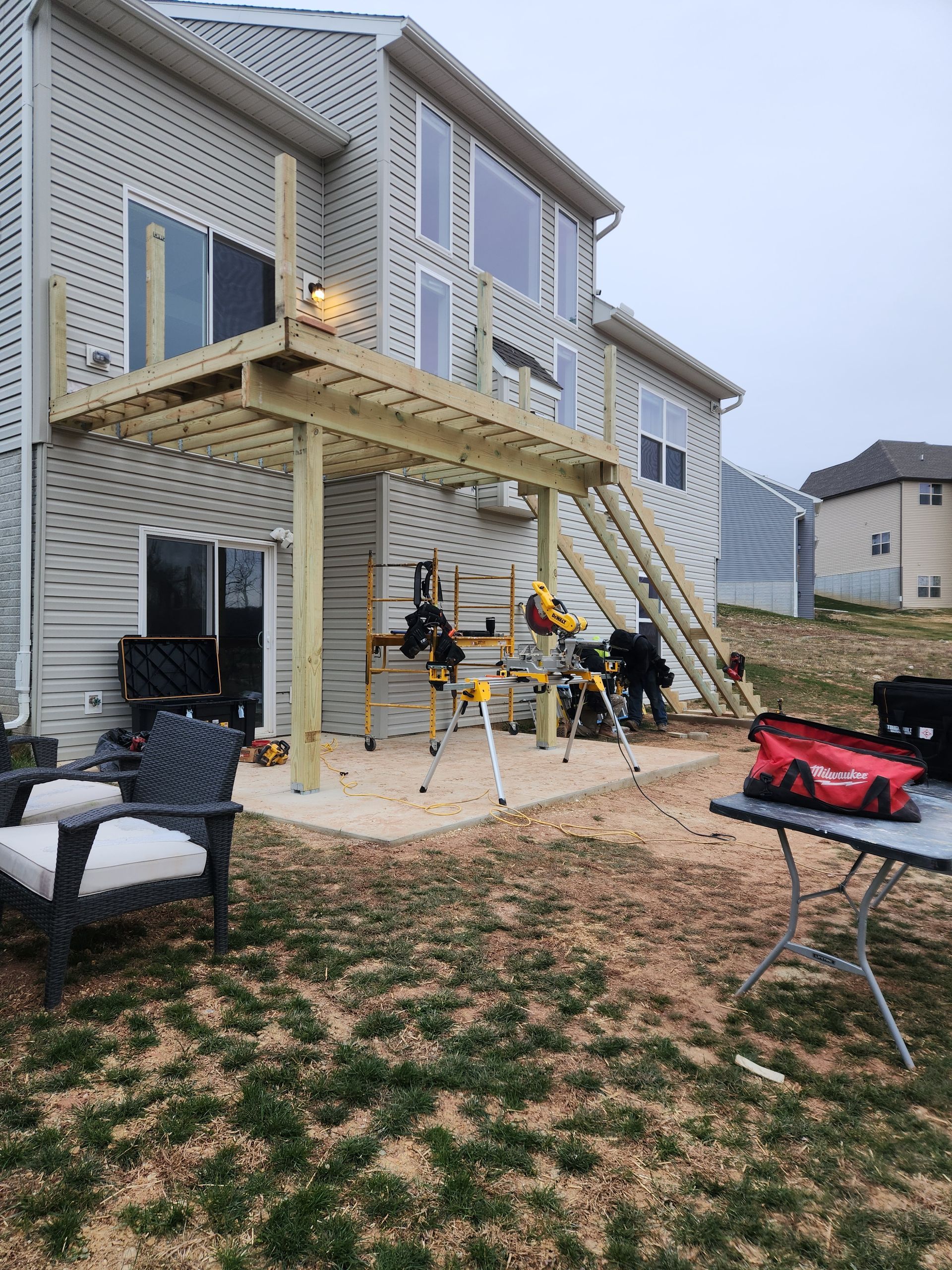 A wooden deck under construction at the back of a beige, two-story house with siding, tools, and a table nearby.