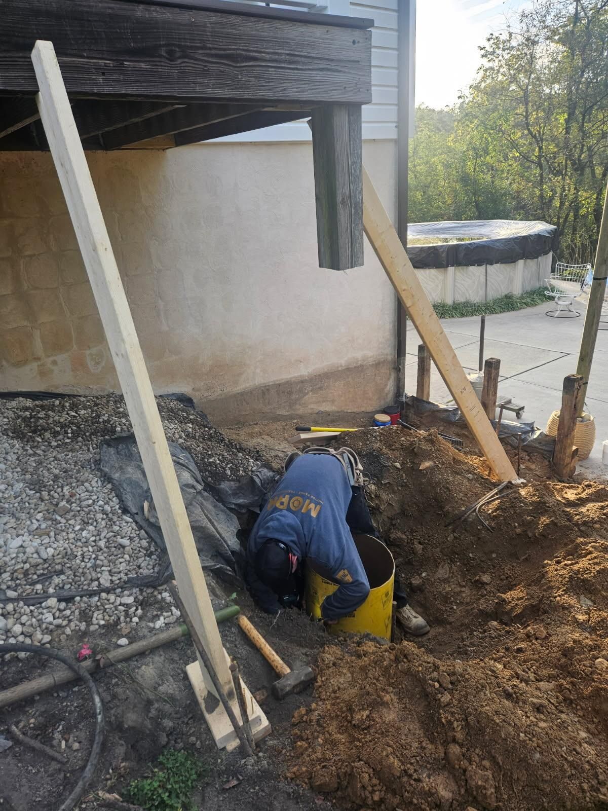 A worker wearing a blue shirt works inside a yellow concrete form tube in an excavated hole beneath a wooden deck.