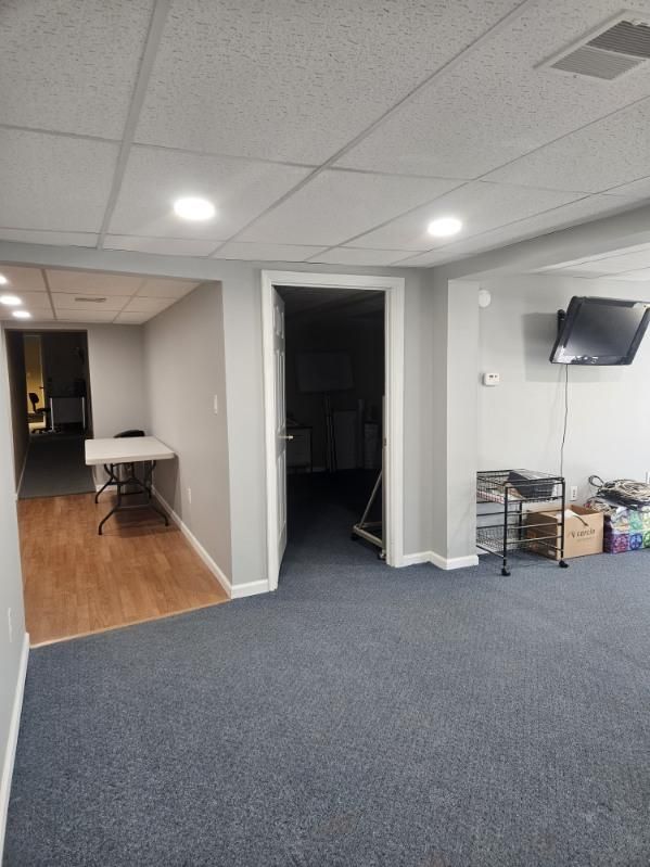 A finished basement with grey carpet, a wood-floored hallway, a table, and a wall-mounted TV.