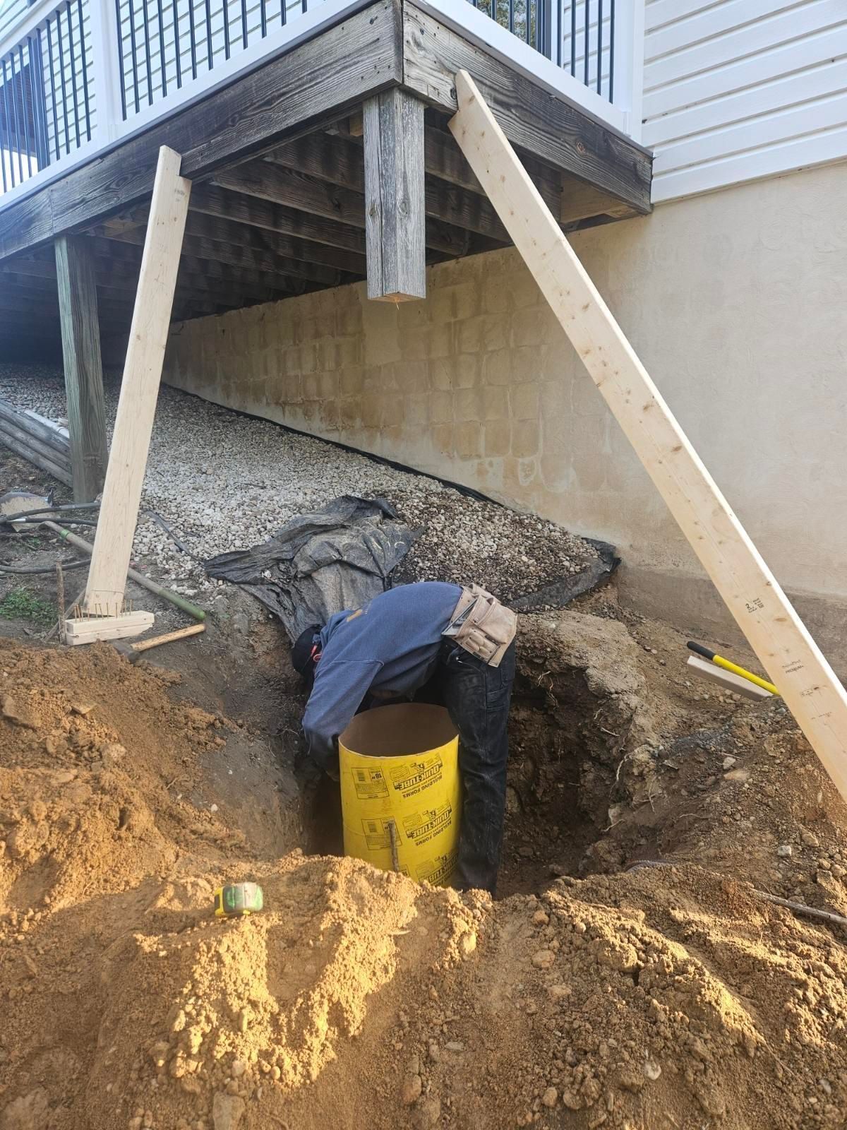 A construction worker installs a yellow cylindrical concrete form in a hole beneath a wooden deck supported by temporary beams.