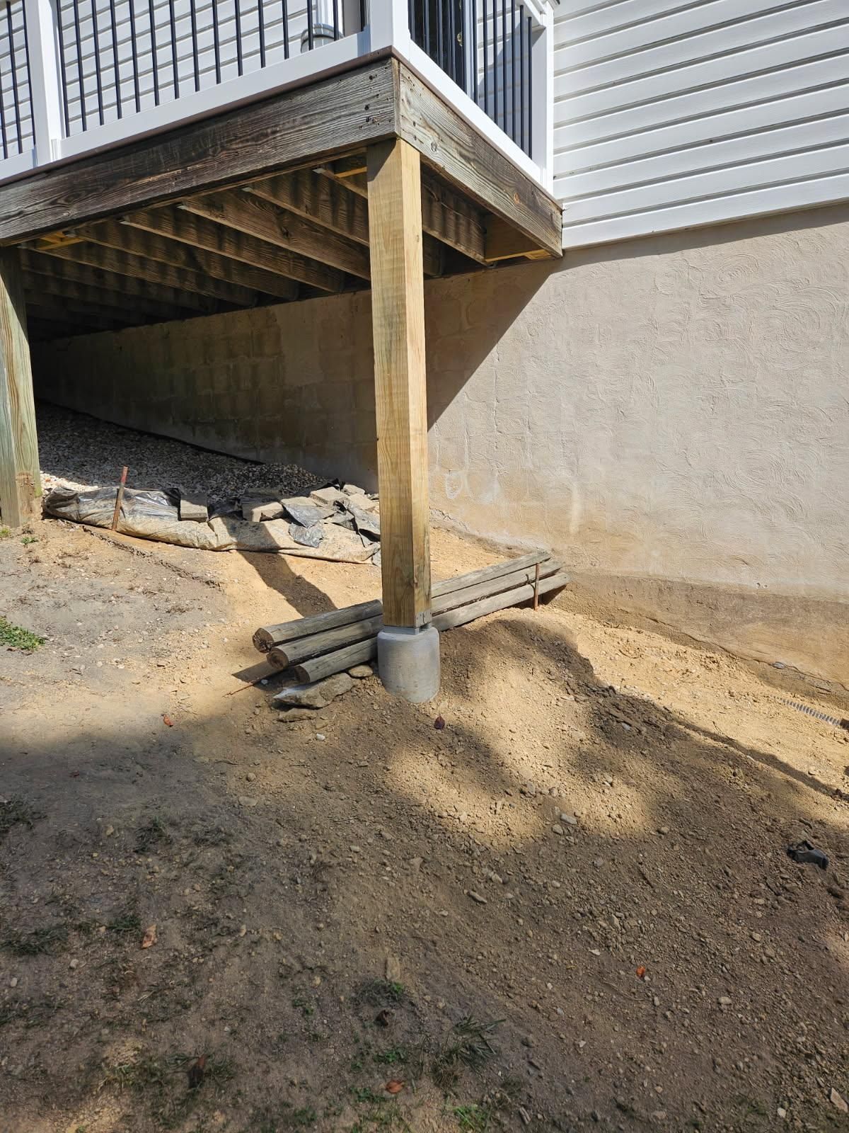 A wooden deck post stands on a concrete footing next to a beige stucco wall, with loose dirt and construction debris below.