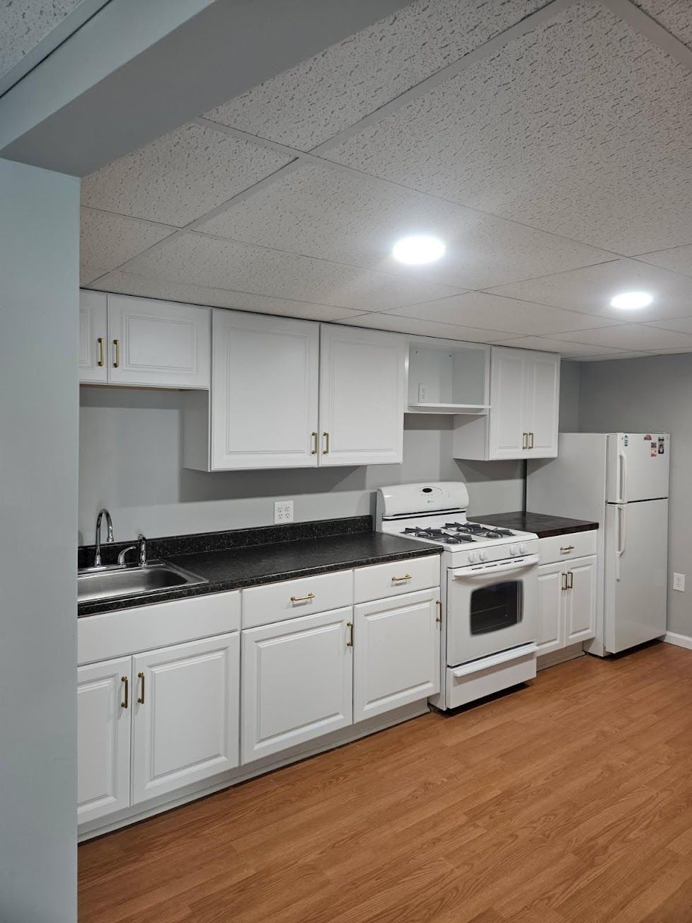 A white kitchen with dark countertops, a gas stove, and a refrigerator against a gray wall, on wood-look flooring.