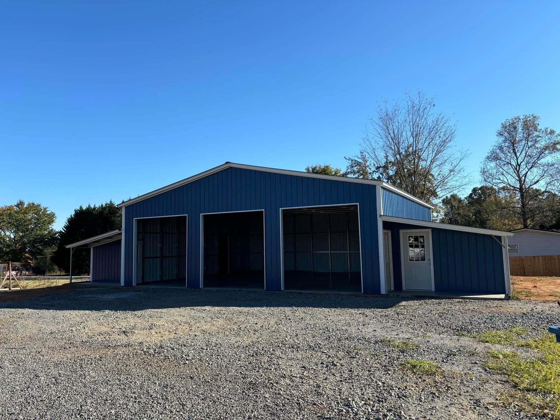 Blue metal building with three garage bays and a single door, set on gravel under a clear sky.