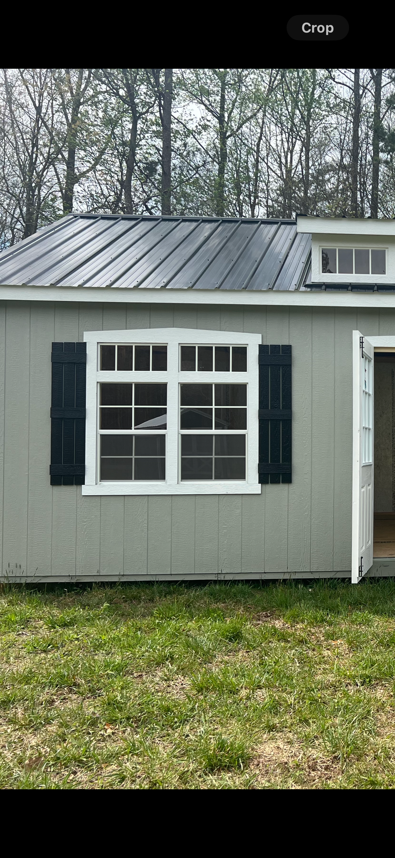A small, gray building with a dark metal roof, white-framed windows, and black shutters, set in a grassy area.