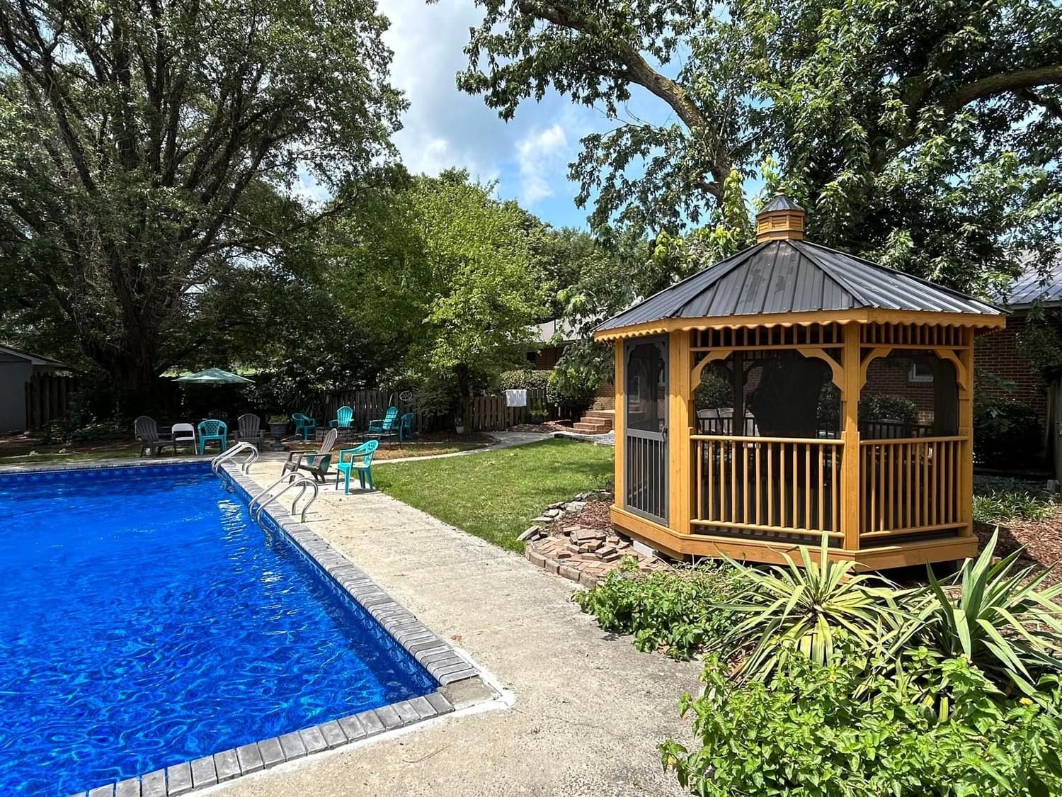 A backyard scene with a blue swimming pool, a wooden gazebo, and green trees under a cloudy sky.