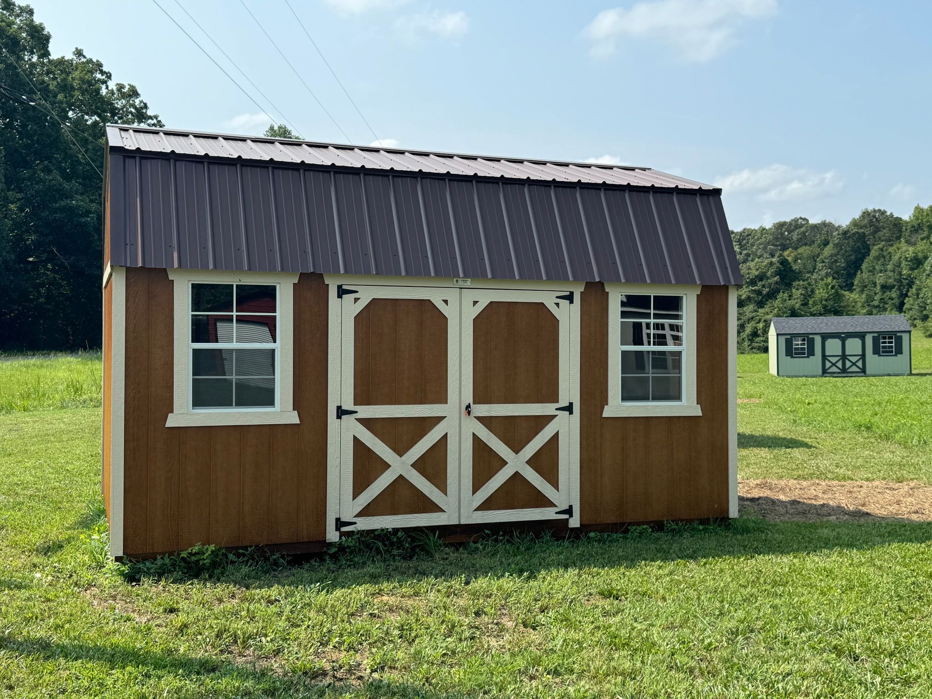 Brown wooden shed with white trim and a brown metal roof, set in a grassy field on a sunny day.
