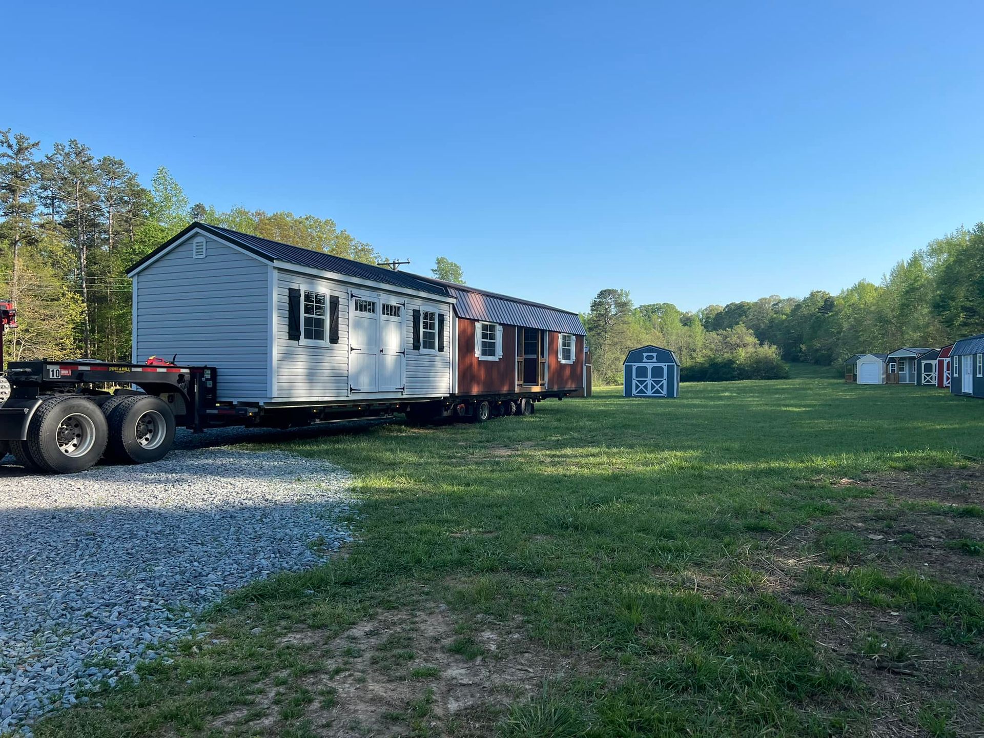 A tiny house on a trailer parked on grass with a gravel driveway, surrounded by trees and other structures.