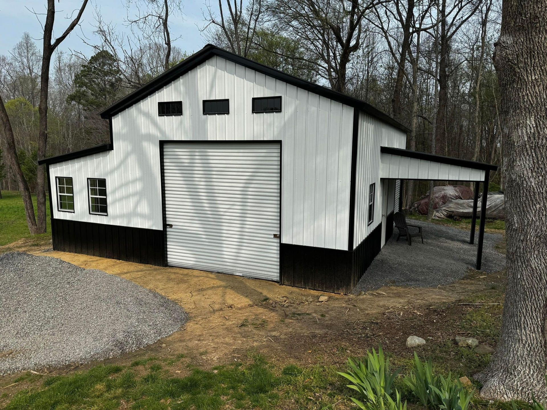 White and black barn with a covered side area, set on a gravel driveway with a wooded background.