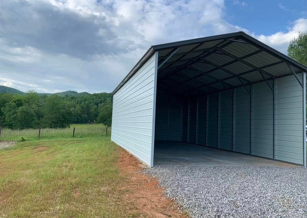 Metal carport with open entrance, gravel floor, light blue siding, and a green landscape backdrop.