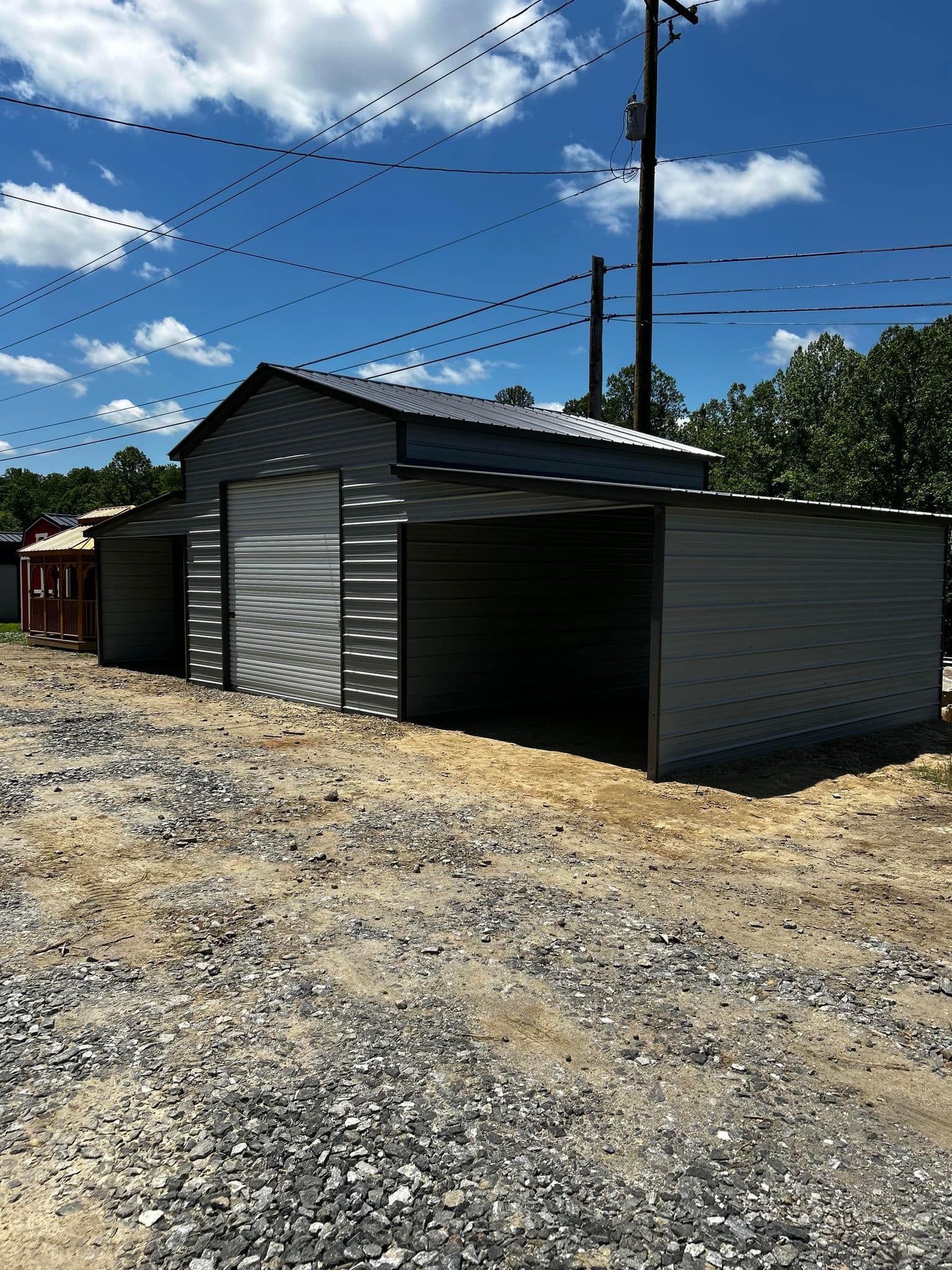 Metal storage sheds on a gravel lot under a blue sky with utility poles and trees in the background.