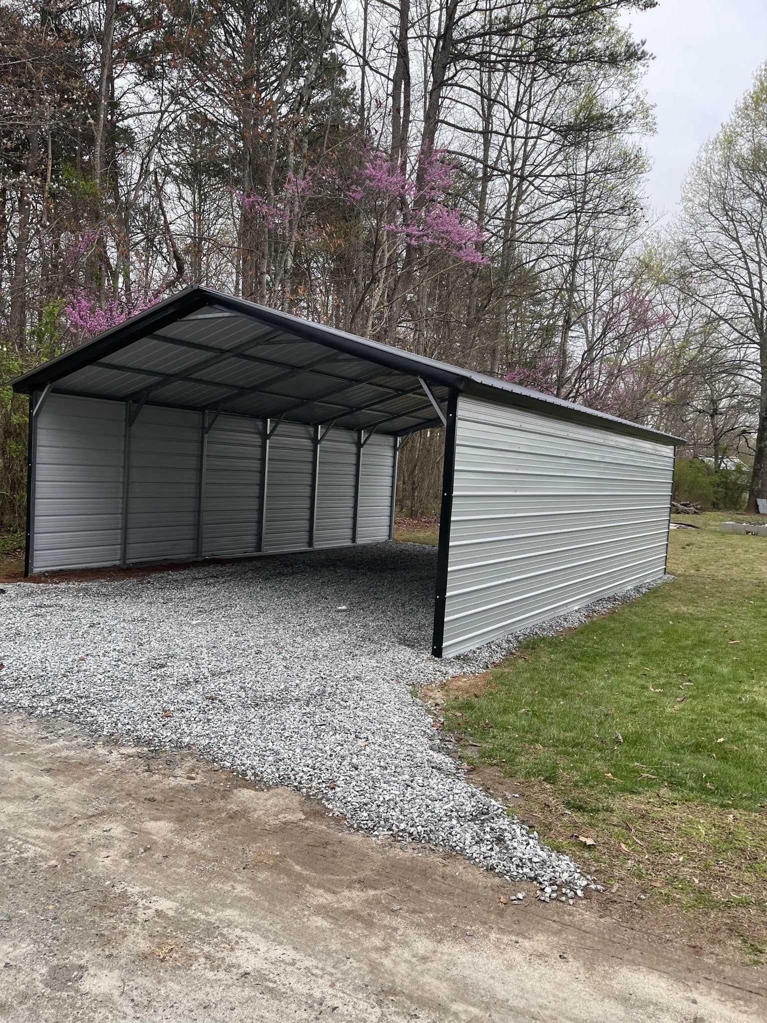Metal carport with a gravel driveway, surrounded by grass and trees. The roof is black, and the structure is light gray.
