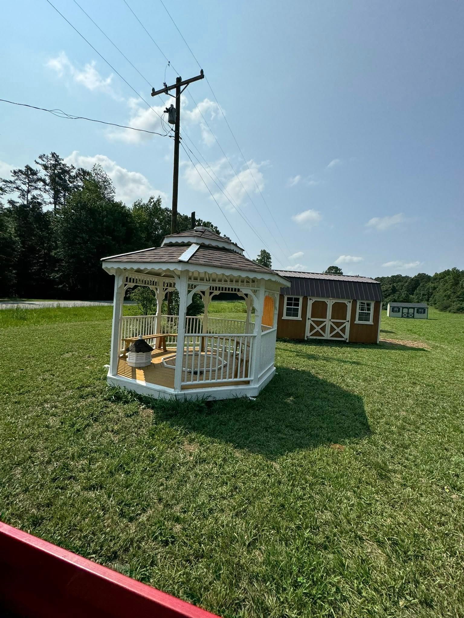 White gazebo in a field, brown building in the background. Green grass, blue sky, and a power line.