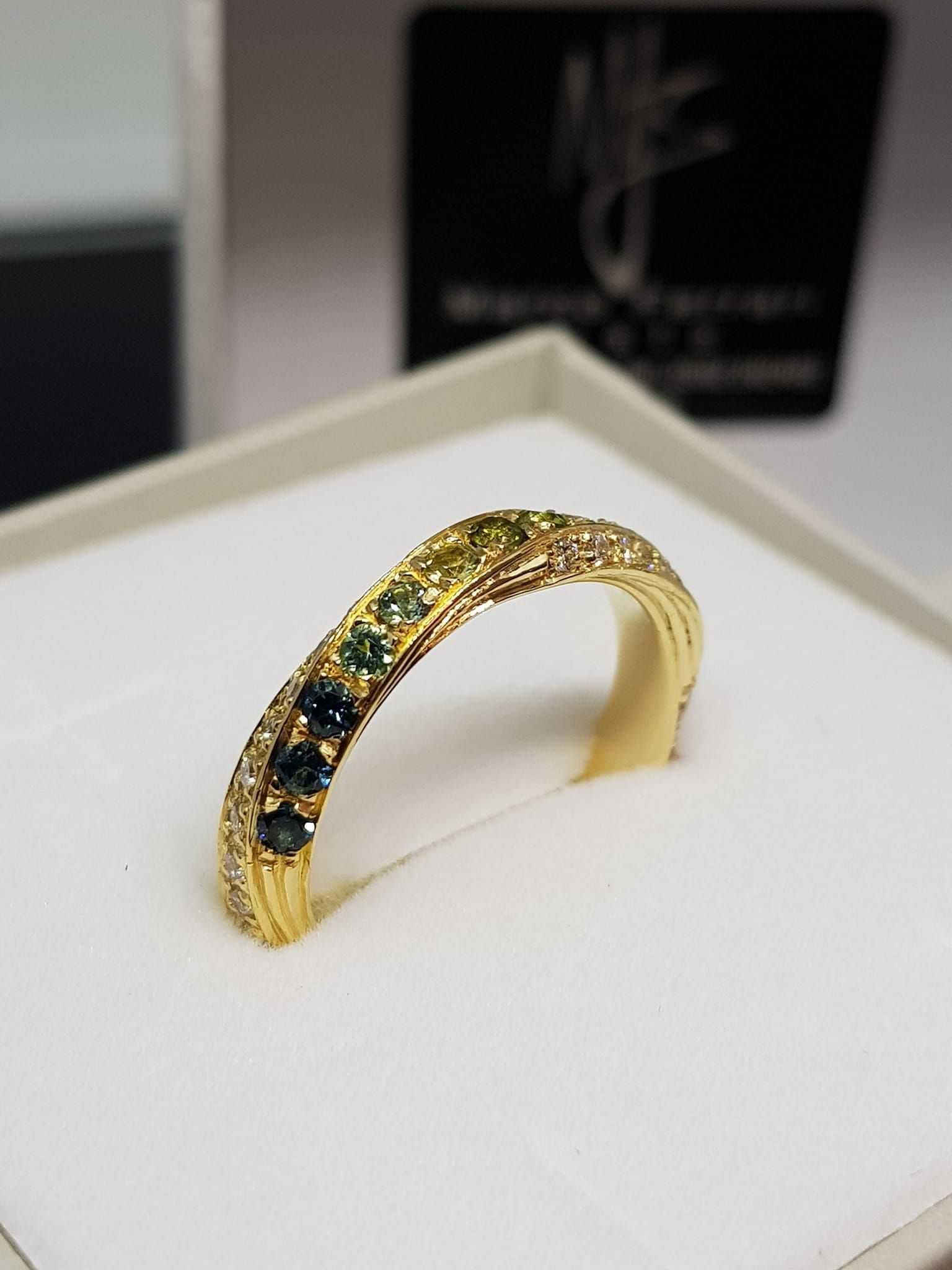 Anello in oro con una fila di gemme verdi e blu, esposto su un supporto bianco all'interno di una scatola.