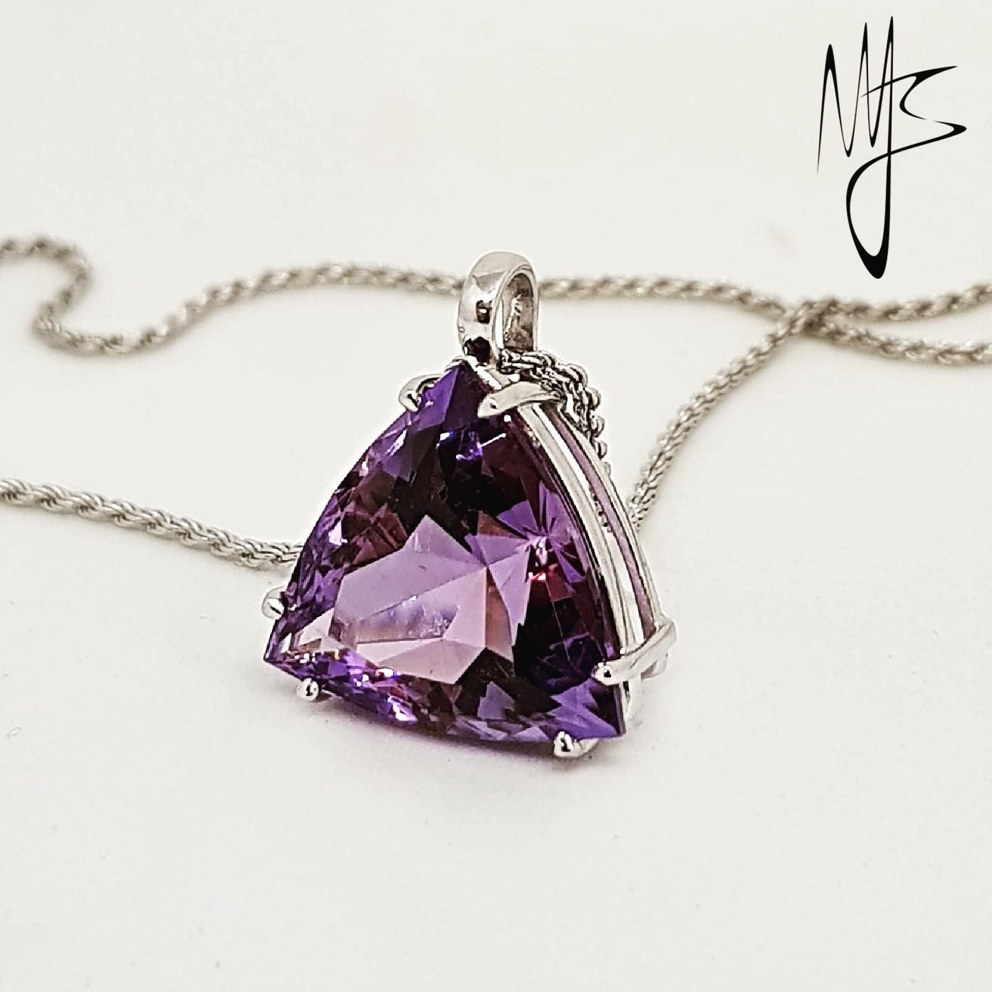 Ciondolo triangolare con pietra preziosa viola su catena d'argento, appoggiato su una superficie bianca.