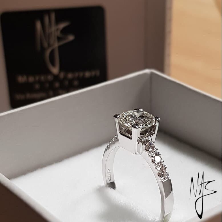 Anello con diamanti in una scatola bianca; fascia in argento con pietre più piccole, grande diamante quadrato centrale.