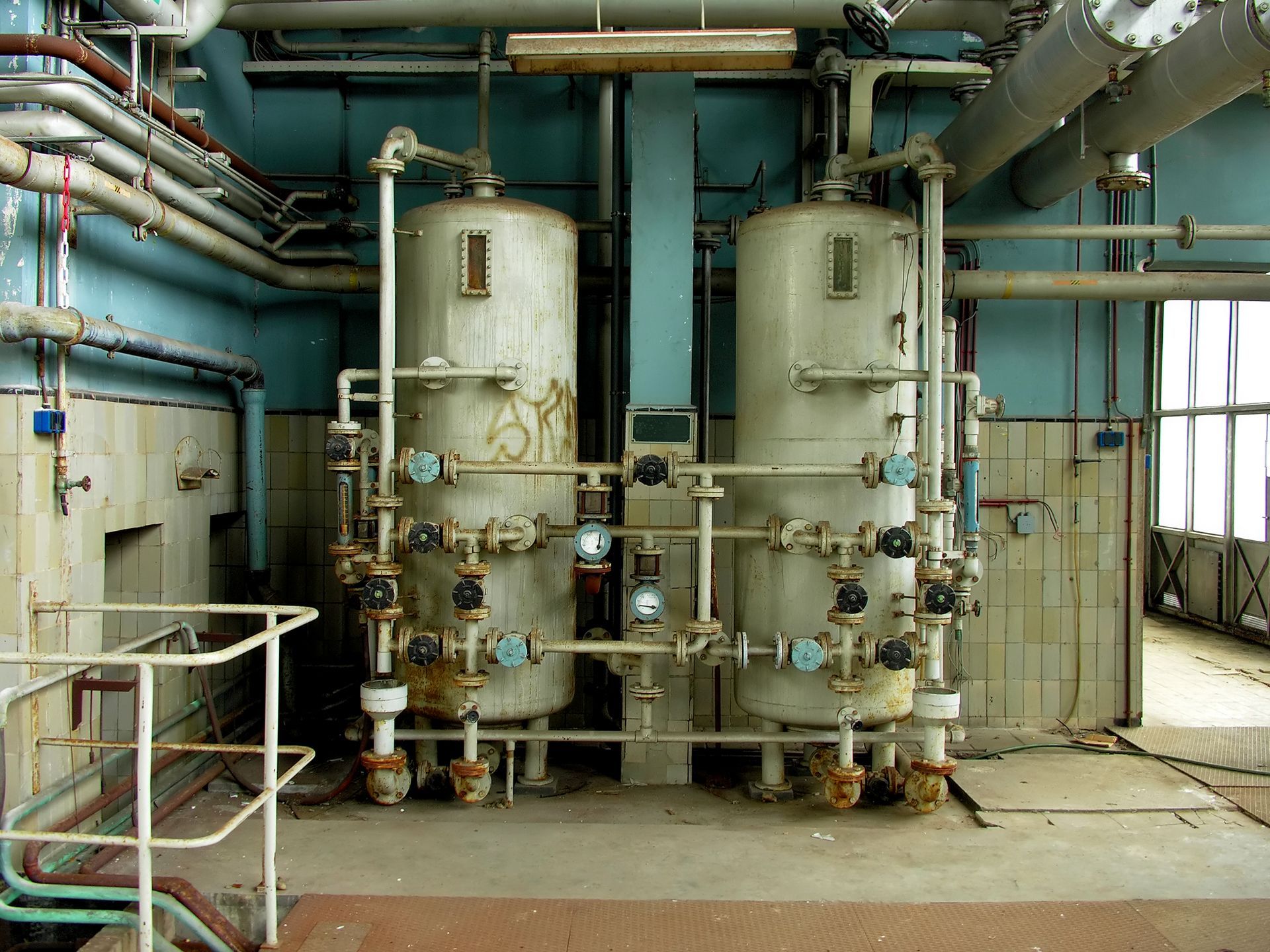 Two large industrial tanks with pipes and valves in a dilapidated, blue-walled room.