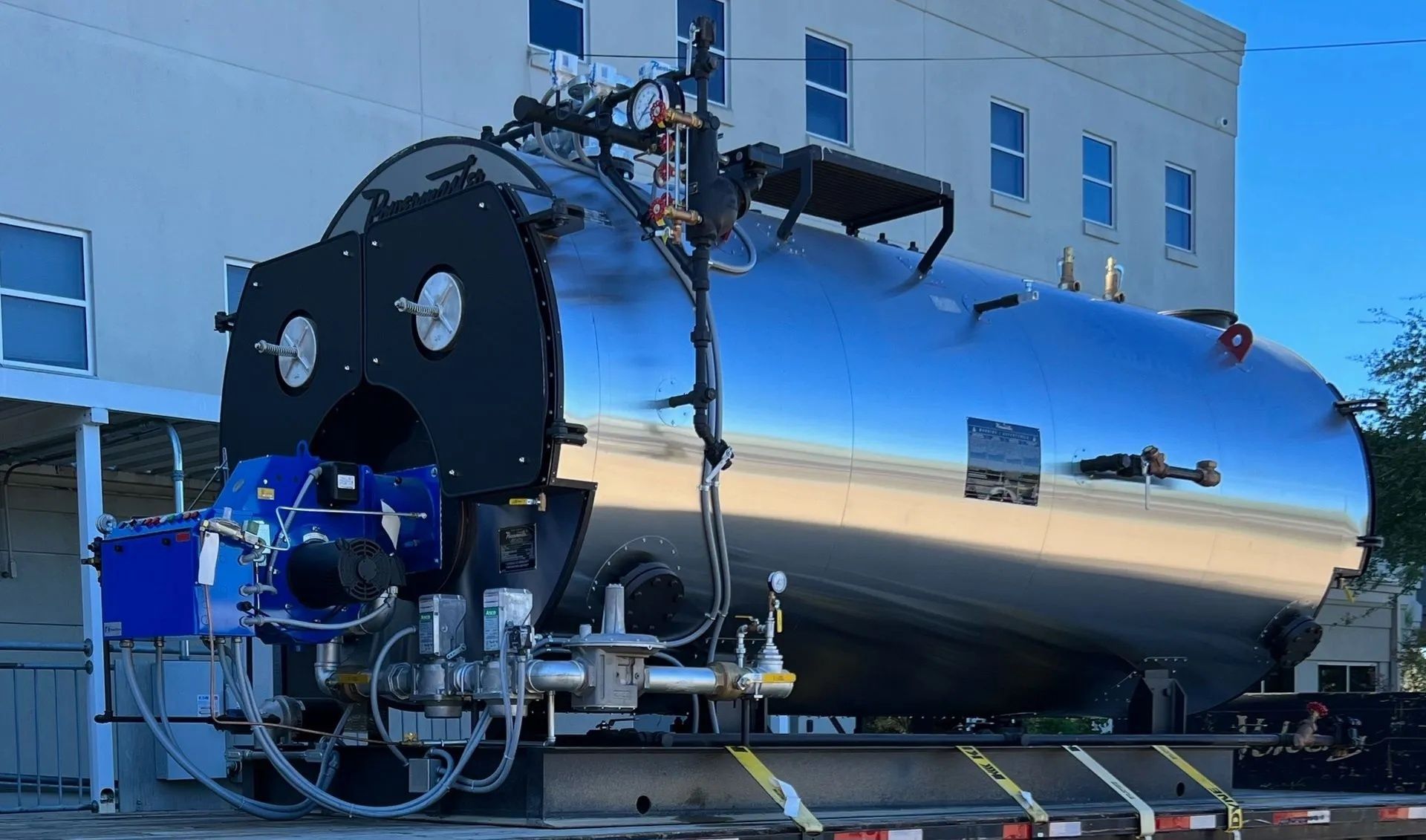 A large, cylindrical industrial boiler with a blue burner on a flatbed truck in front of a building.
