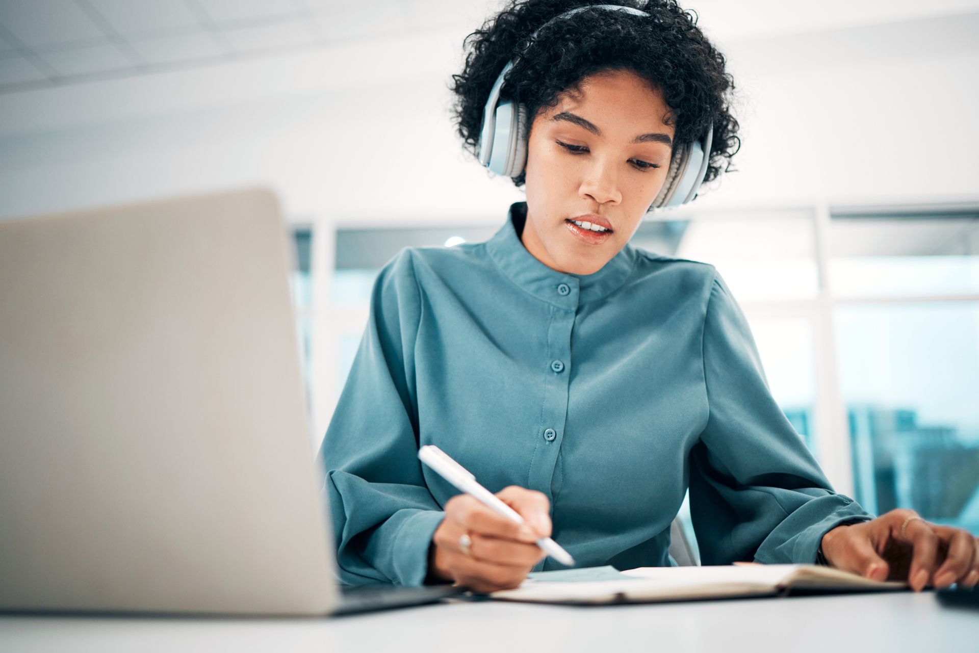 A woman wearing headphones is writing in a notebook while using a laptop computer.