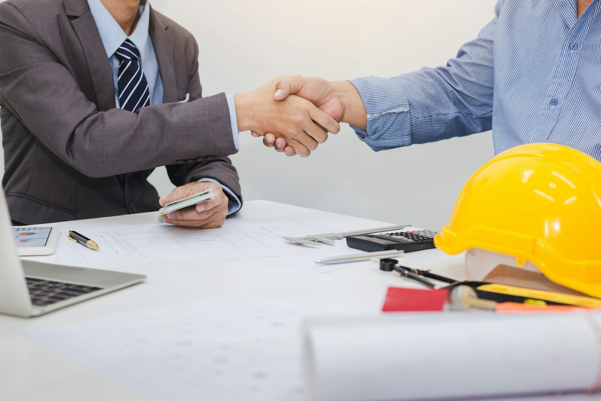 Two people shaking hands over a desk with documents, laptop, and yellow hard hat