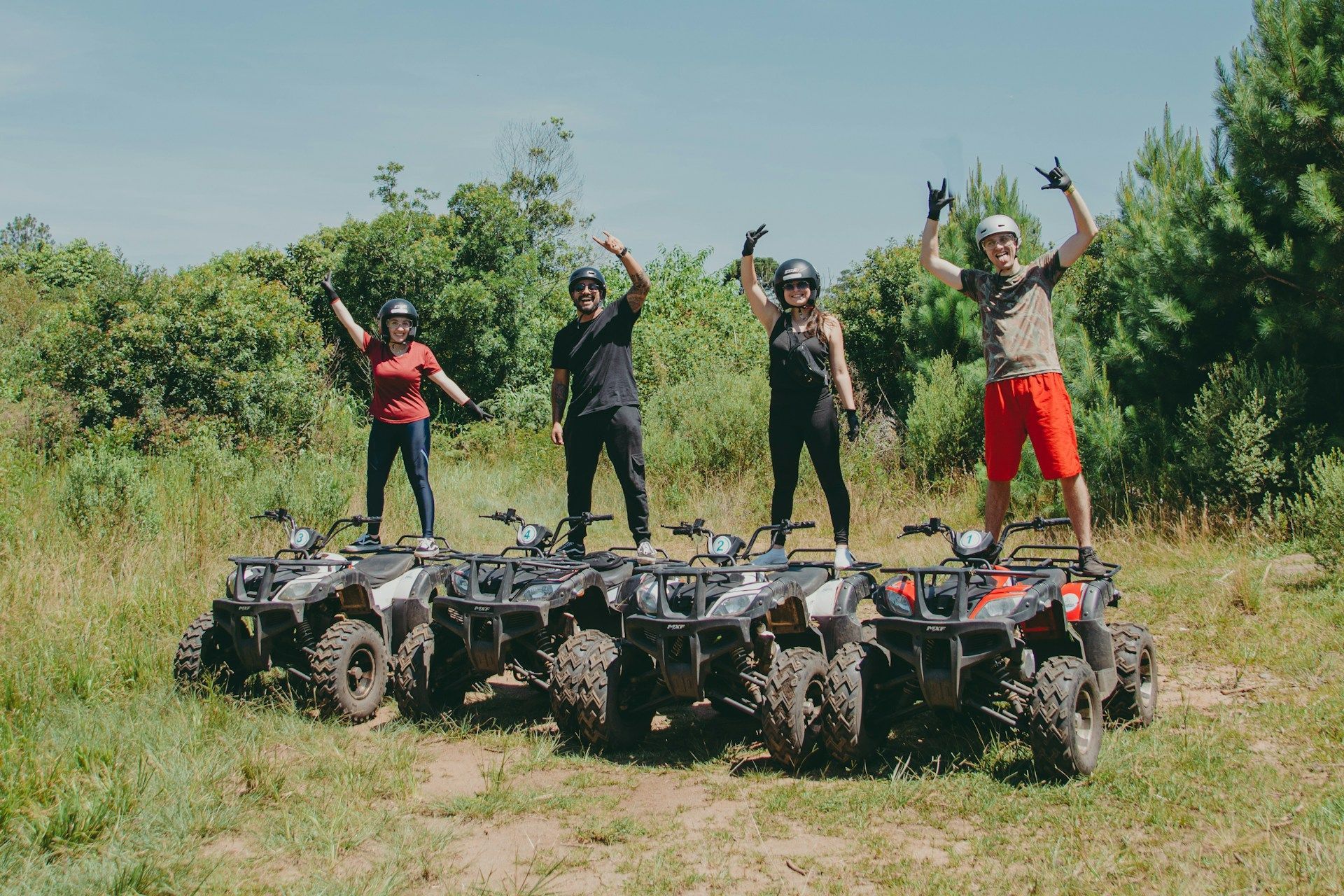 Four people standing on four ATVs in a grassy field, cheering with arms raised.
